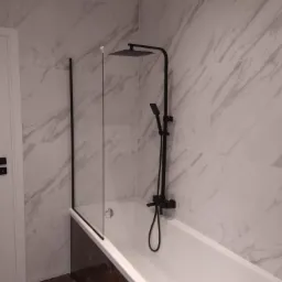 Nowoczesna łazienka z białą wanną, czarną armaturą i szklanym parawanem, ściany wyłożone płytkami imitującymi marmur.