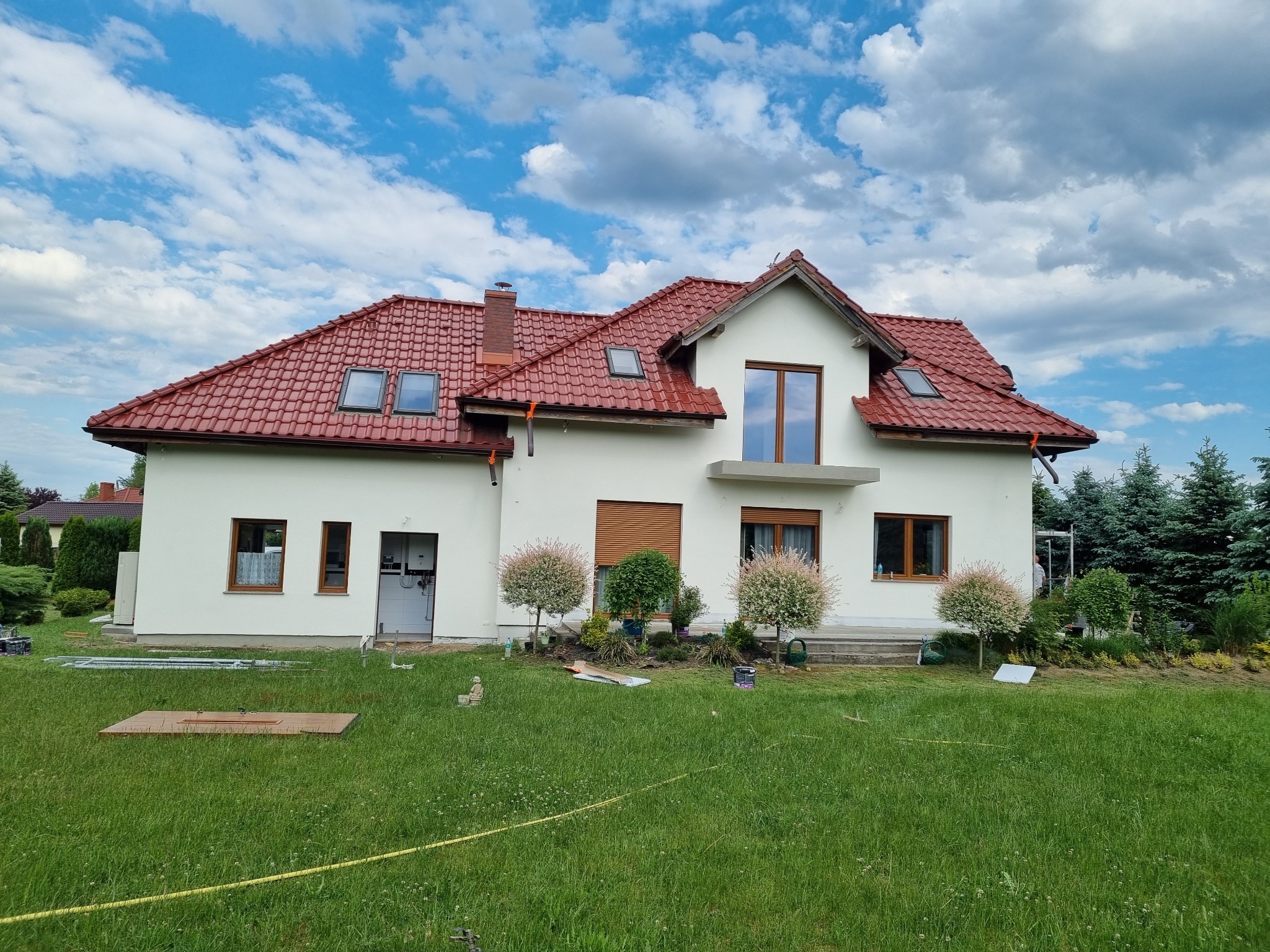 Dom jednorodzinny we Wrocławiu z jasną elewacją i czerwoną dachówką, widok z trawnika. Na elewacji widoczne świeże ślady malowania.