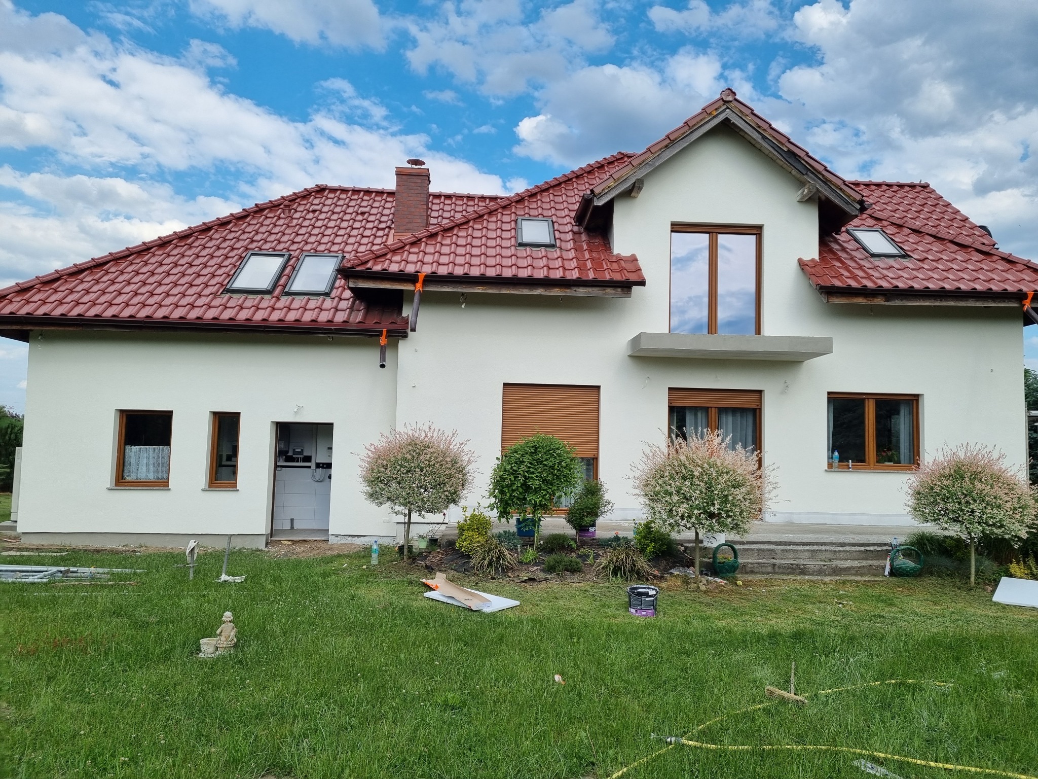 Świeżo otynkowany dom jednorodzinny z czerwoną dachówką, widoczne okna dachowe, elewacja w kolorze złamanej bieli, dekoracyjny ogródek z młodymi drzewkami i rozłożony sprzęt budowlany na trawniku.