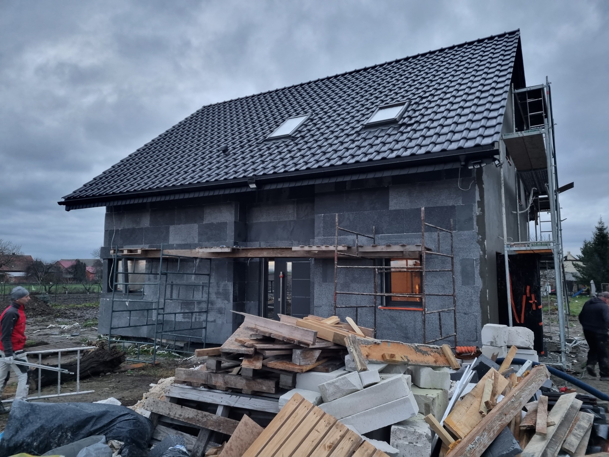Dom w trakcie ocieplania ścian zewnętrznych, widoczne rusztowania, stos materiałów budowlanych, ciemna elewacja, okna dachowe i pochmurne niebo.