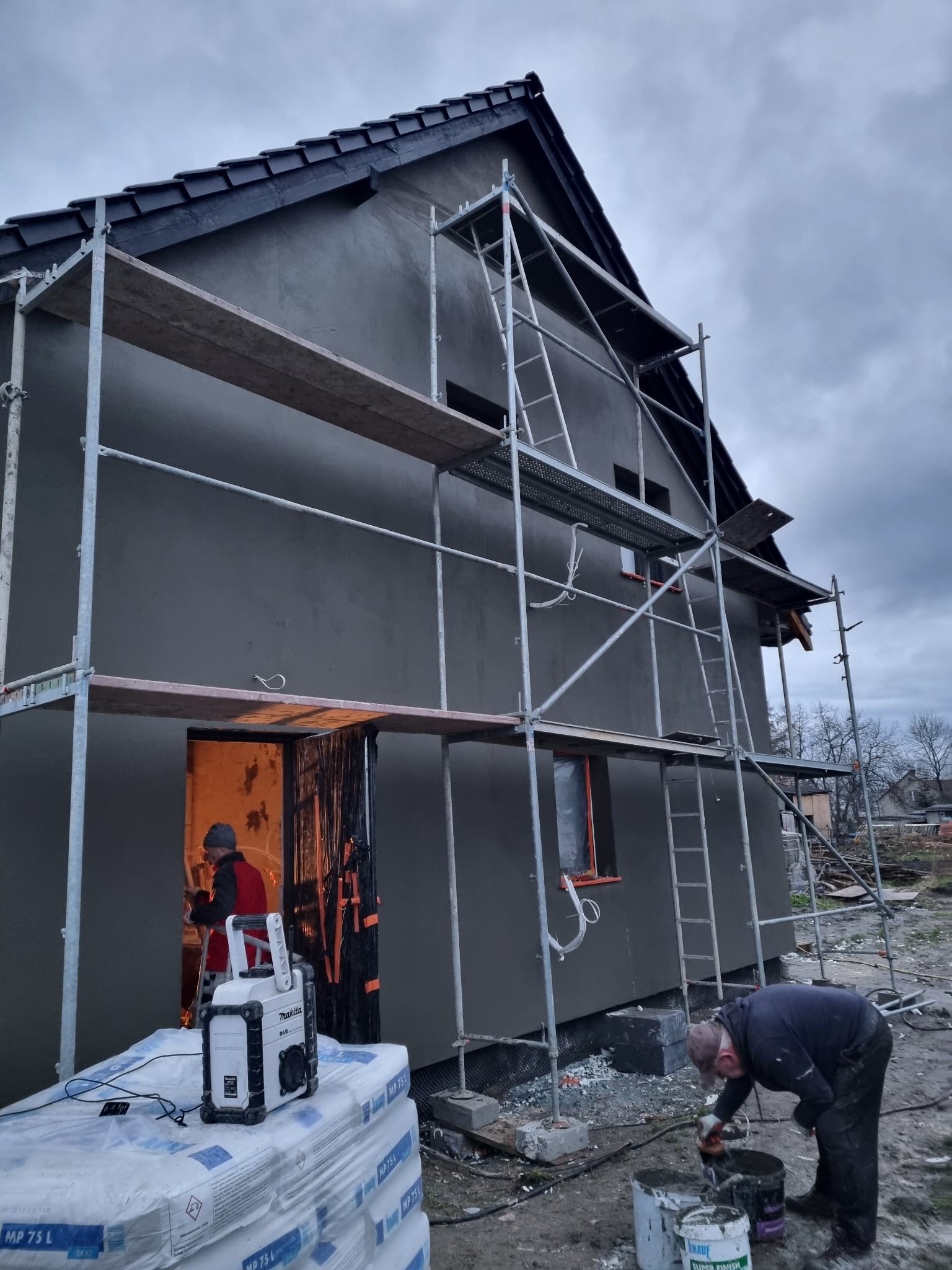 Elewacja domu w trakcie szpachlowania, widoczne rusztowania, pracownik mieszający zaprawę, radio budowlane Makita na workach zaprawy, pochmurne niebo.