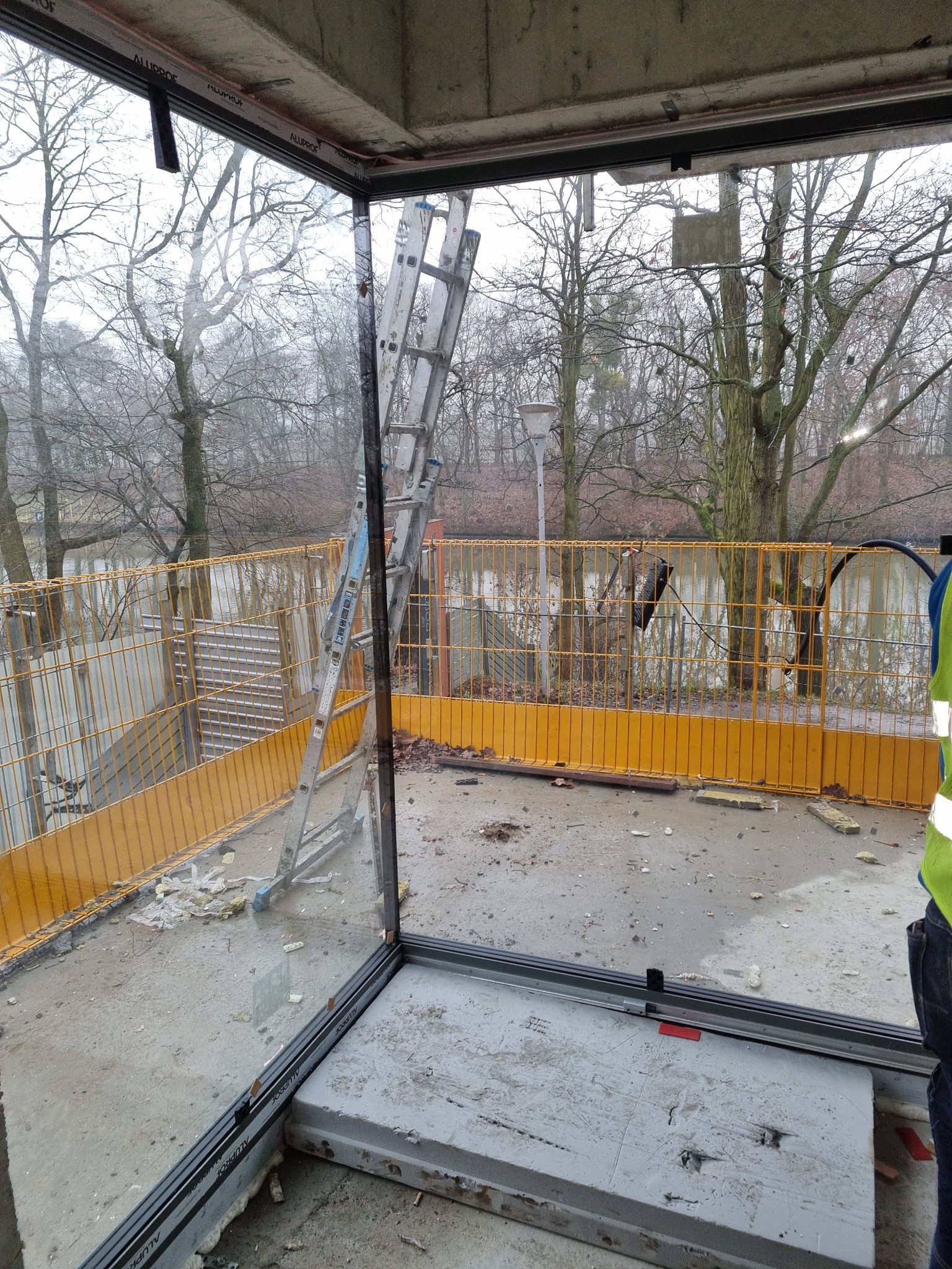 Surowy stan budowy: montaż okien z widokiem na park, drabina oparta o ramę okienną, fragment żółtego ogrodzenia ochronnego w tle, widoczne ślady prac budowlanych na podłodze.