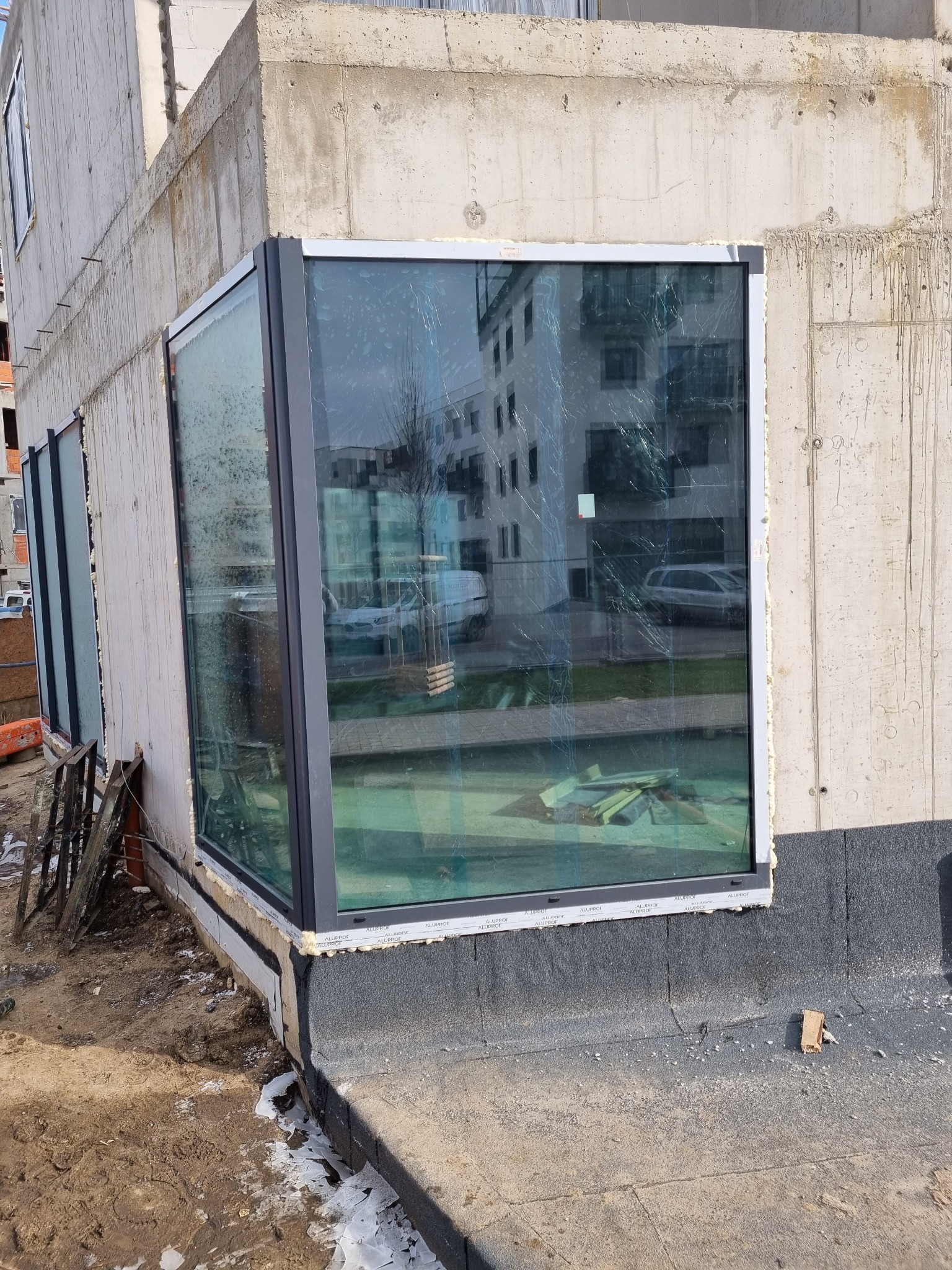 Narożne okno z ciemną ramą zamontowane w surowym budynku w trakcie budowy, widoczne ślady prac murarskich i ziemia wokół.