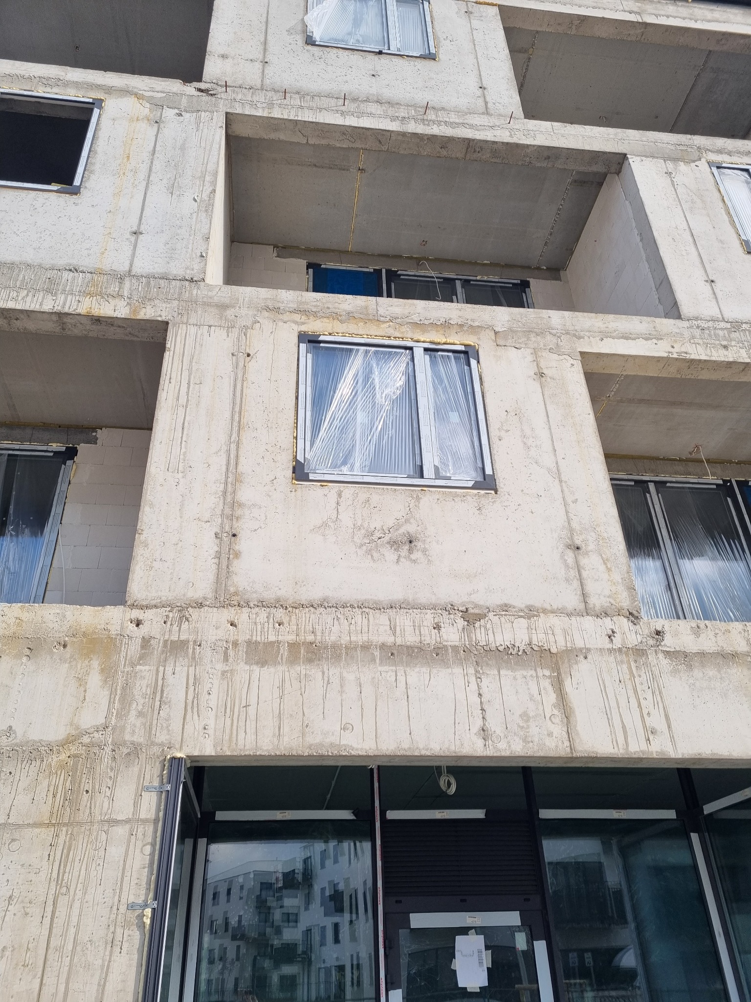 Surowy betonowy budynek w trakcie budowy, z zamontowanymi oknami zabezpieczonymi folią ochronną, widoczne ślady po zalewaniu betonu.