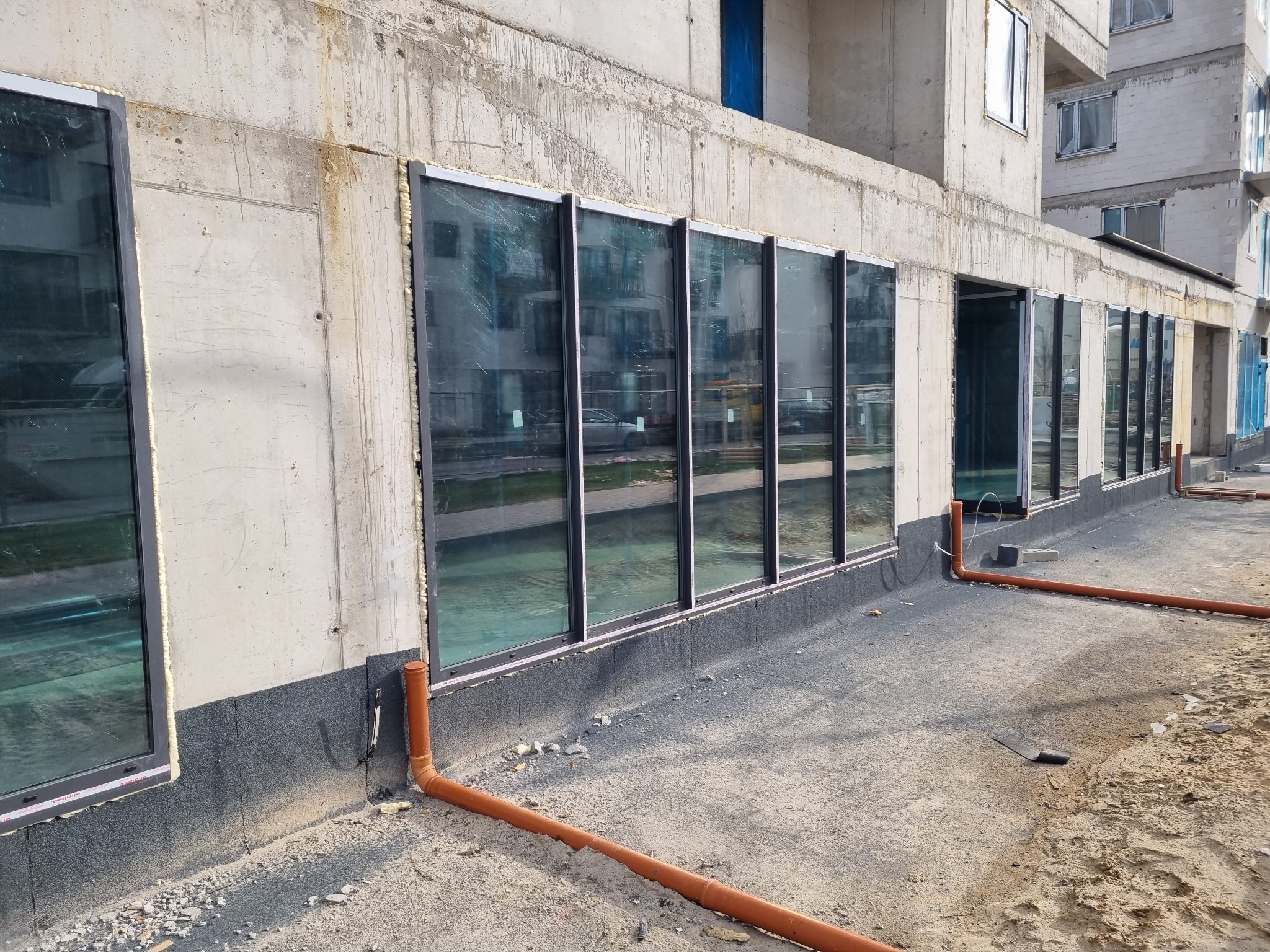 Montaż okien w surowym budynku z widoczną izolacją fundamentów i rurami odprowadzającymi wodę, pianka montażowa wokół ram okiennych.