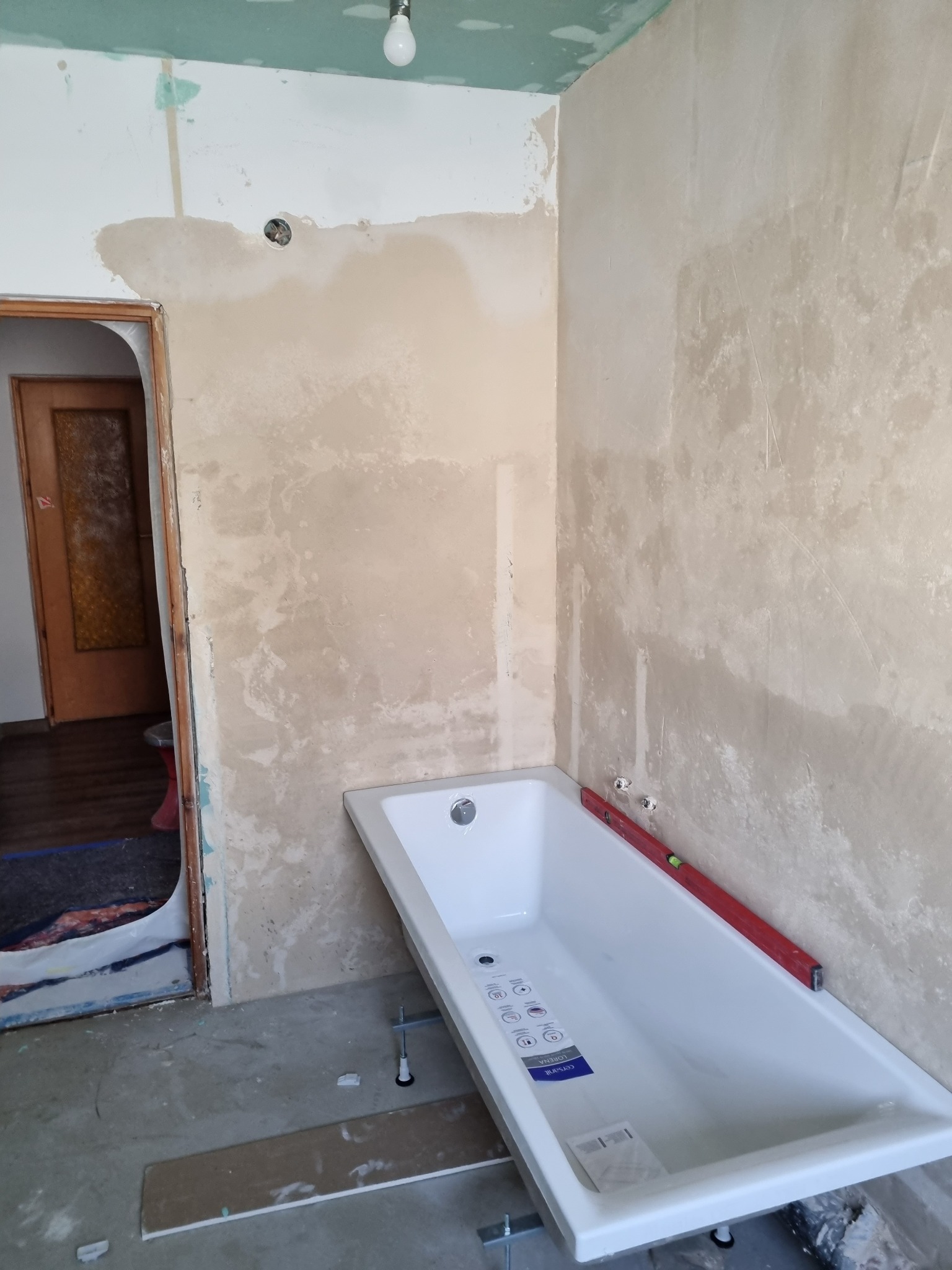 Biała wanna ustawiona w surowym stanie łazienki, z widocznymi śladami po gładzi na ścianach i poziomnicą, sugerująca etap przygotowania do dalszych prac wykończeniowych.