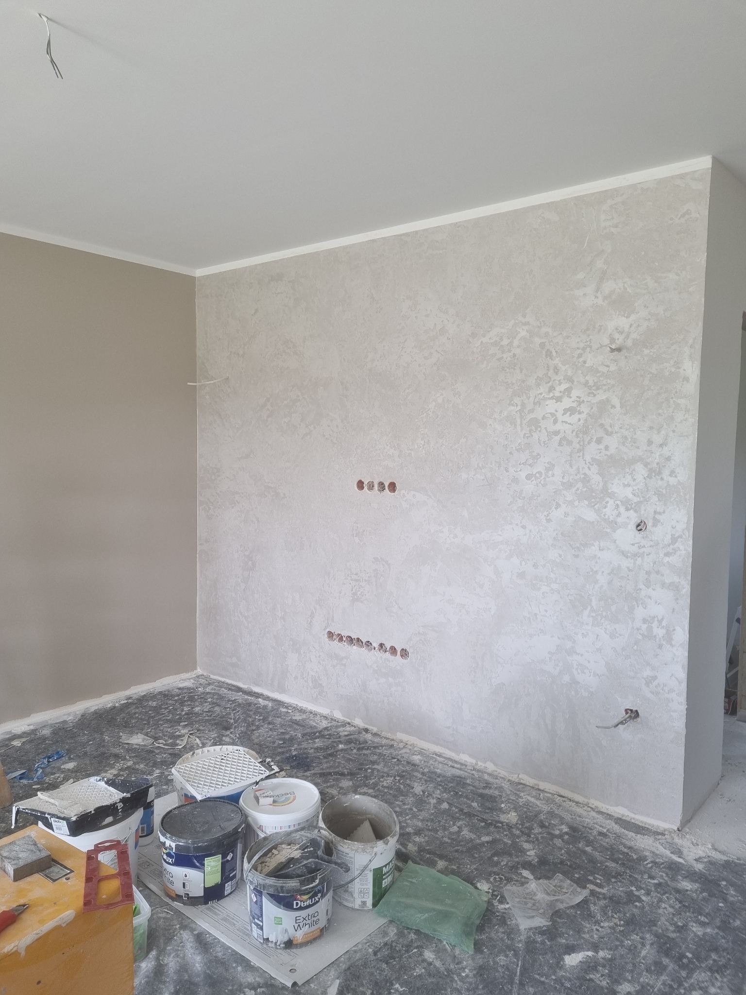 Pokój w trakcie malowania: jedna ściana beżowa, druga z dekoracyjną fakturą w odcieniach szarości, widoczne puszki z farbą Dulux Extra White i narzędzia malarskie na podłodze zabezpieczonej folią.