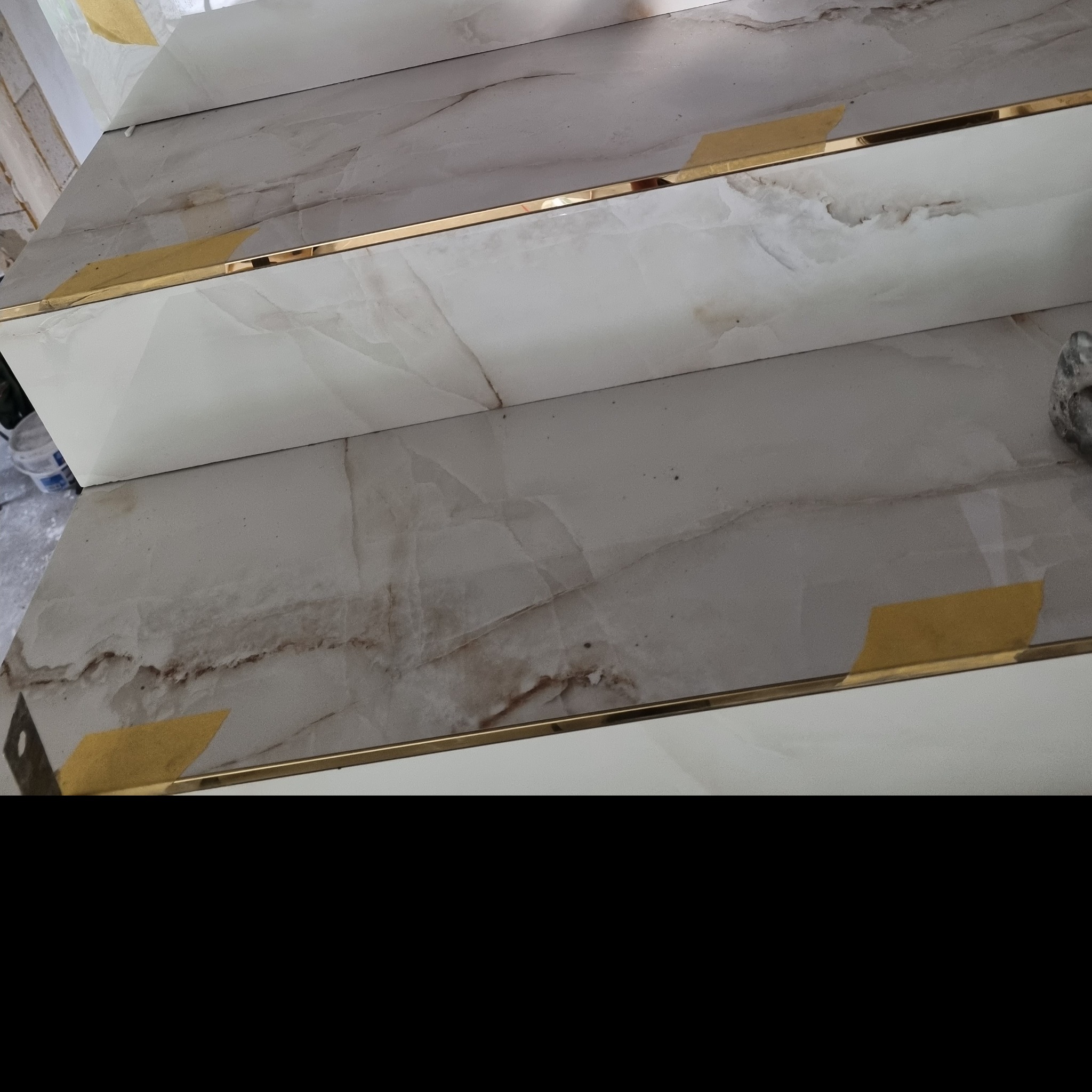 Fragment wykończonych schodów z płytkami imitującymi marmur, zabezpieczonych żółtą taśmą malarską i wykończonych złotymi listwami krawędziowymi.