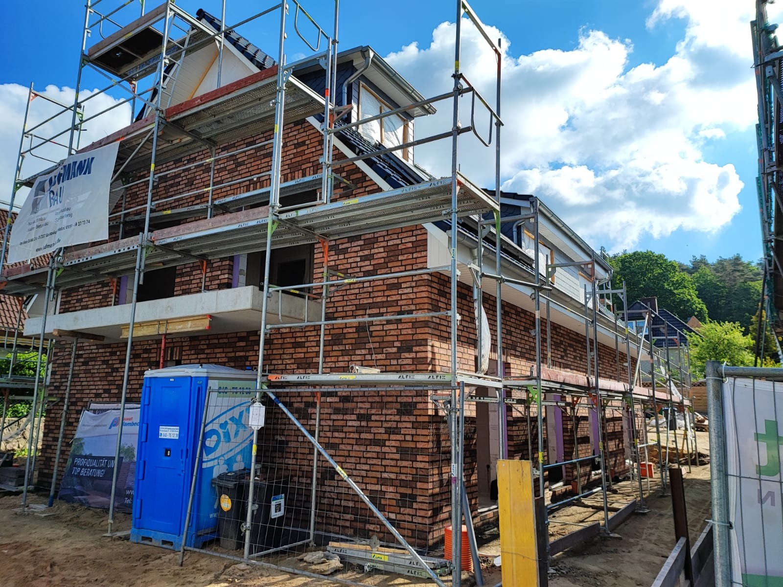 Budowa domu z czerwonej cegły w trakcie realizacji, widoczne rusztowania, przenośna toaleta i materiały budowlane.
