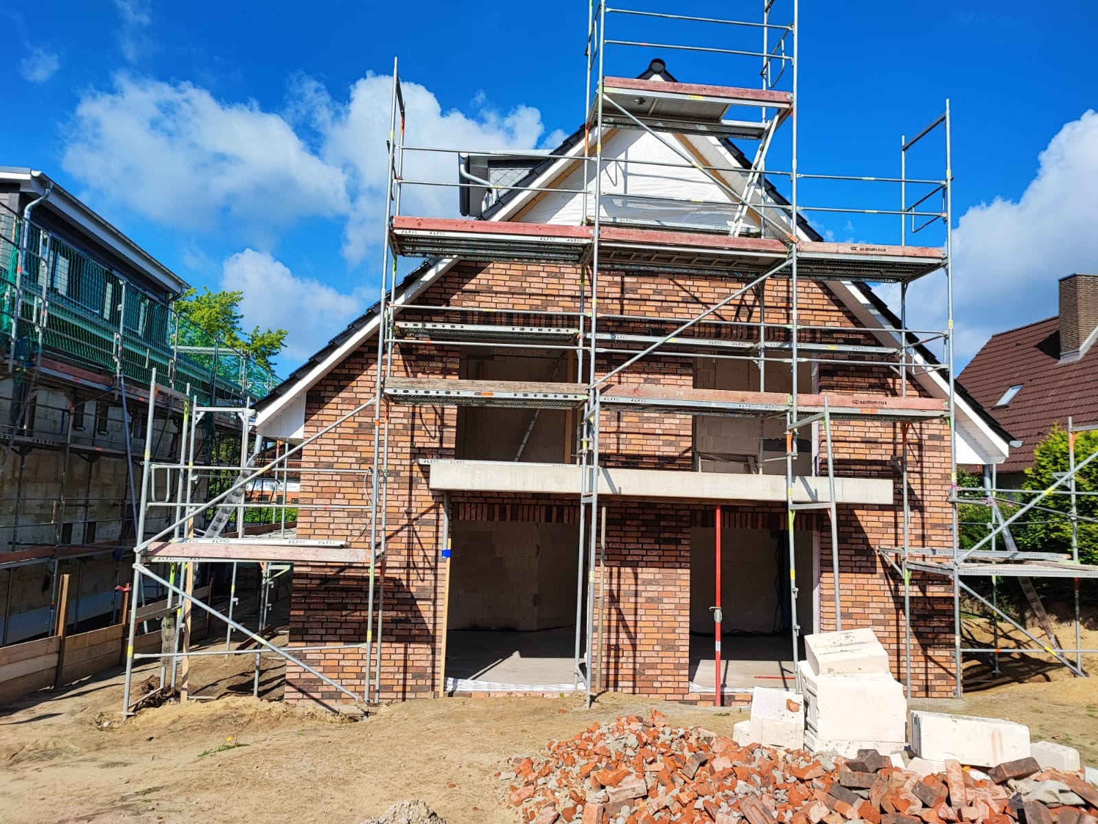 Budynek w trakcie murowania z cegły, otoczony rusztowaniami, z widocznym stosem cegieł na pierwszym planie i błękitnym niebem w tle.