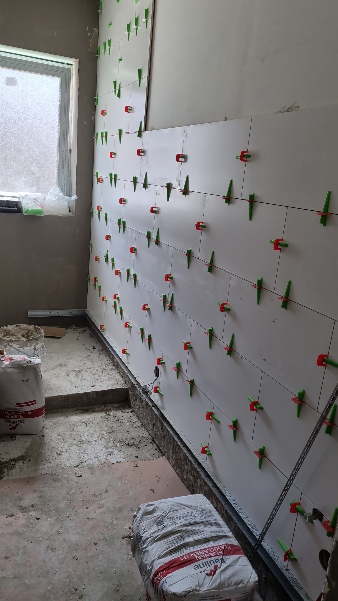 Układanie białych płytek na ścianie z użyciem systemu poziomowania płytek z zielonymi i czerwonymi klipsami, widoczne worki z klejem do płytek i okno w tle.