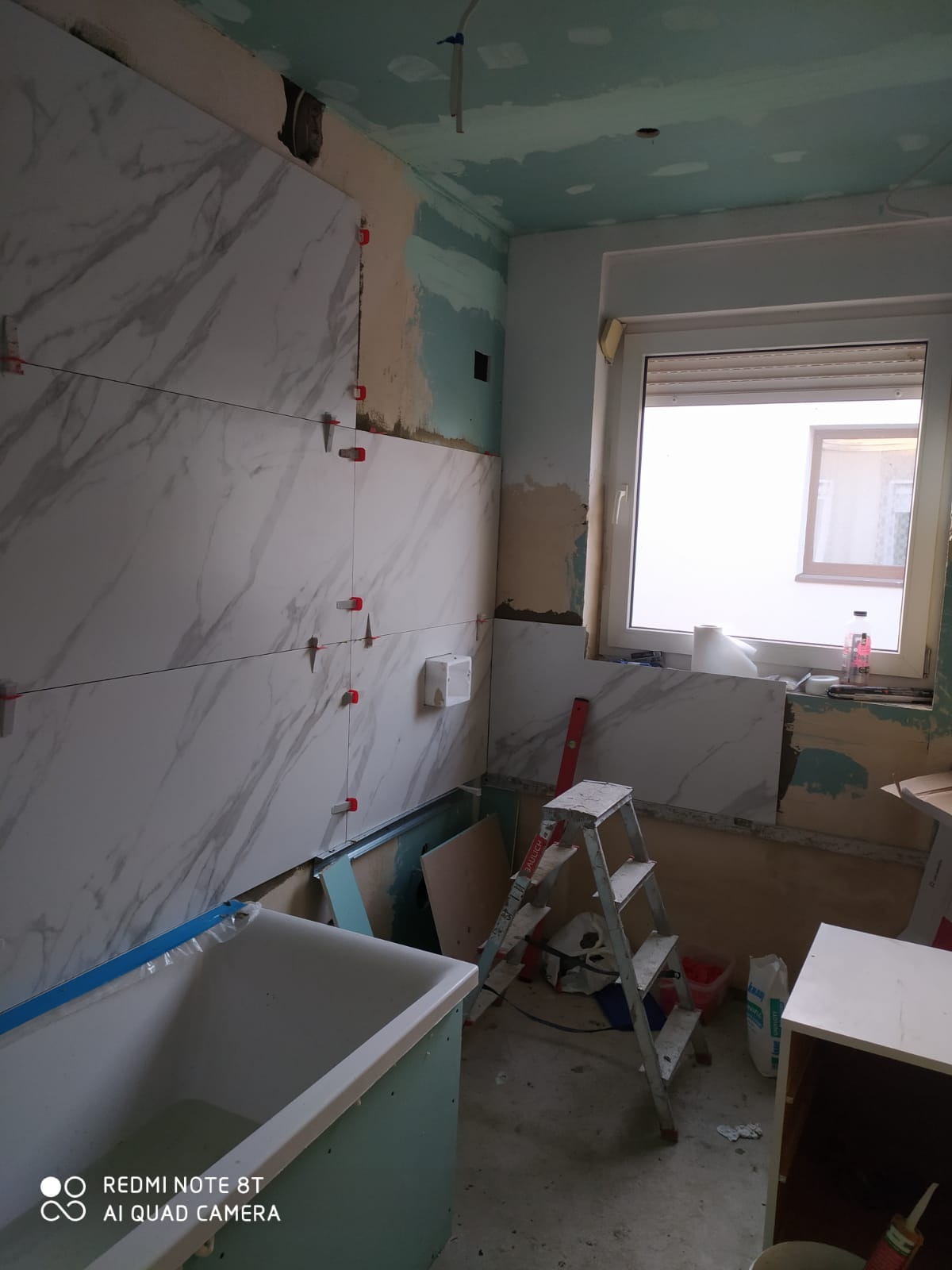 Układanie płytek imitujących marmur na ścianie w łazience w trakcie remontu, widoczne poziomice, drabina, wanna w obudowie z zielonych płyt gipsowych, okno z roletami i niedokończone ściany.