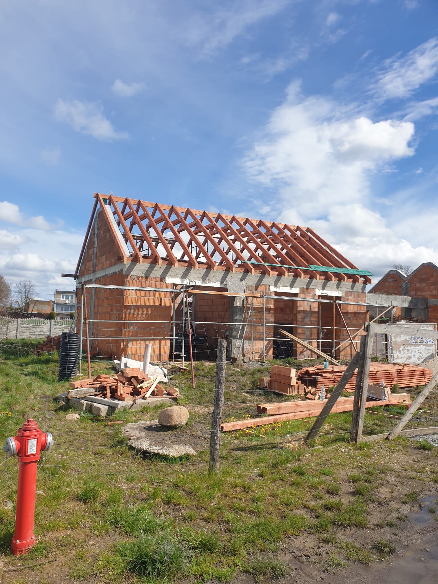 Budowa domu jednorodzinnego w stanie surowym otwartym, widoczne ściany z czerwonej cegły, więźba dachowa, materiały budowlane na działce, hydrant przeciwpożarowy w tle błękitne niebo z chmurami.