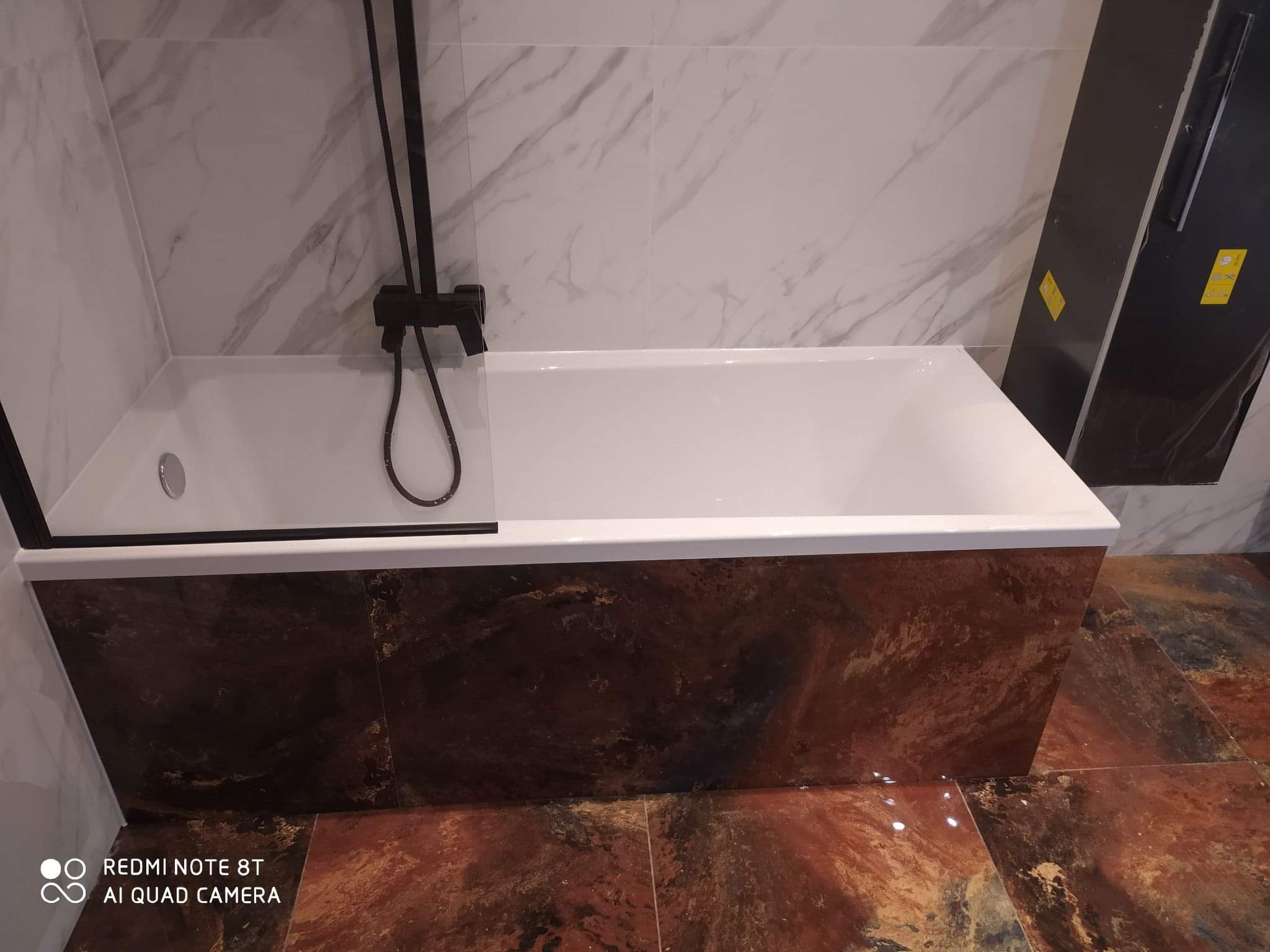 Nowoczesna wanna z czarną armaturą, obudowana płytkami imitującymi rdzę, na tle marmurowych płytek ściennych i podłogi w podobnej tonacji.