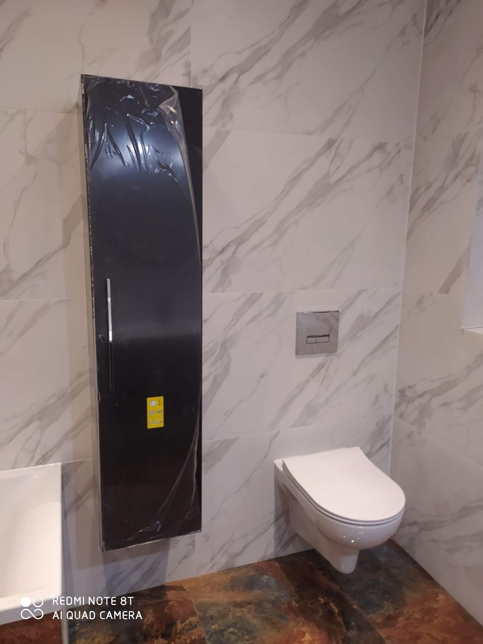 Nowoczesna łazienka z marmurowymi płytkami, wisząca toaleta i czarna szafka w folii ochronnej przed montażem, widoczny fragment umywalki.