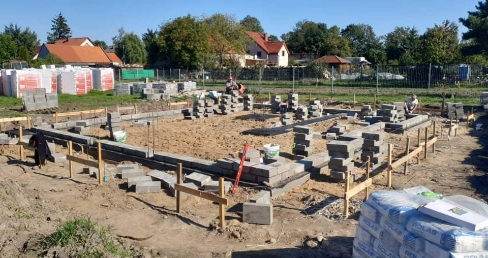 Wykop pod fundamenty z ułożonym szalunkiem i częściowo wymurowanymi bloczkami betonowymi, widoczne zapasy bloczków i pracownicy w oddali.