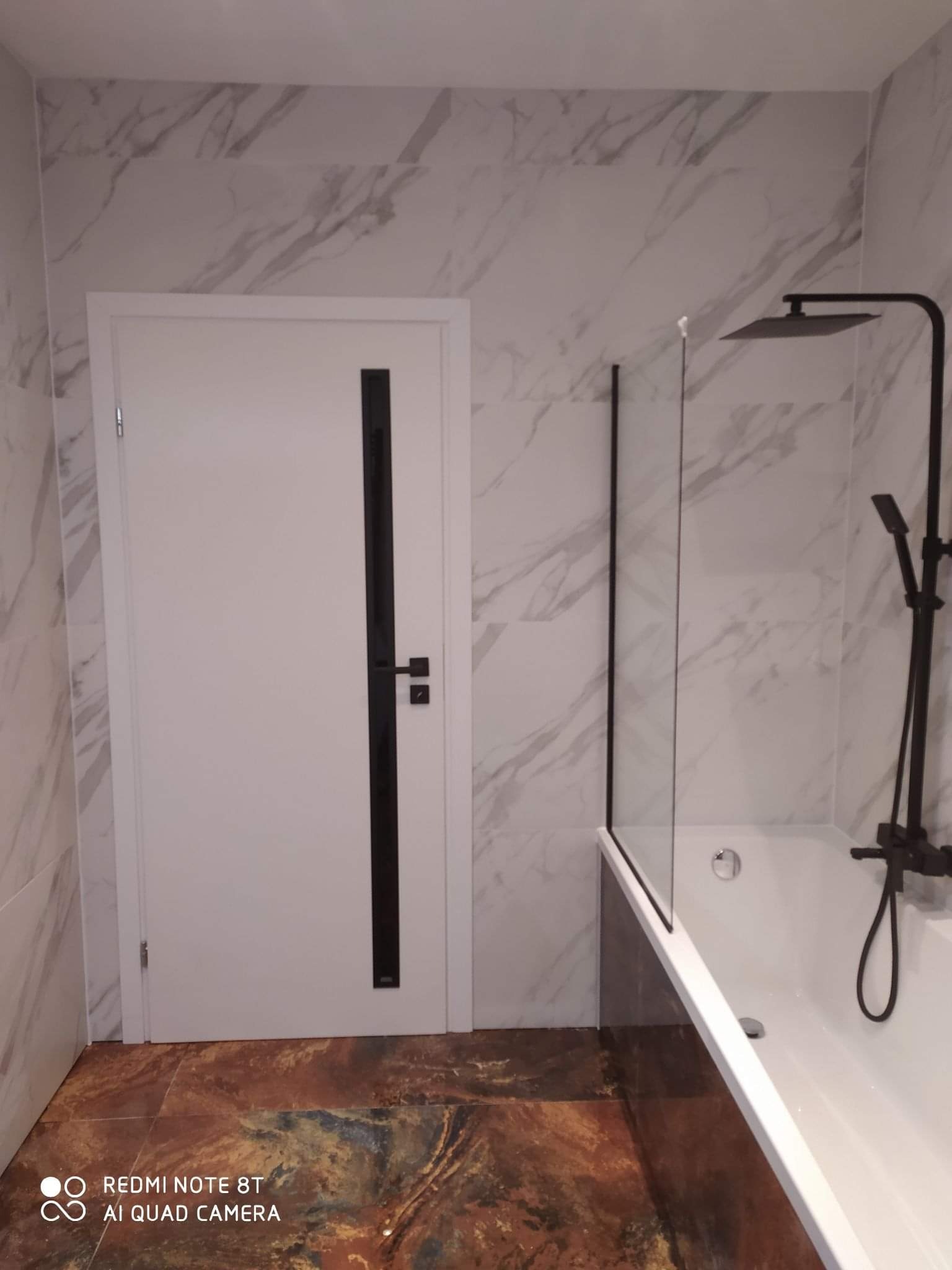 Wykończona łazienka z białą wanną z czarną armaturą, szklanym parawanem, białą stolarką drzwiową z czarnymi akcentami i marmurowymi płytkami ściennymi.