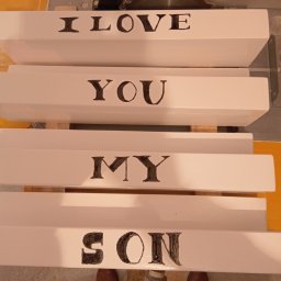 Miko wood - Cztery białe, lakierowane deski z ręcznie malowanymi czarnymi literami układającymi się w napis 'I LOVE YOU MY SON', leżące na stole warsztatowym obok piły tarczowej.