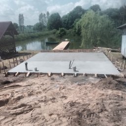 Miko wood - Świeżo wylana, betonowa płyta fundamentowa z wystającymi rurami instalacyjnymi, otoczona drewnianym szalunkiem na tle malowniczego krajobrazu z jeziorem i pomostem.