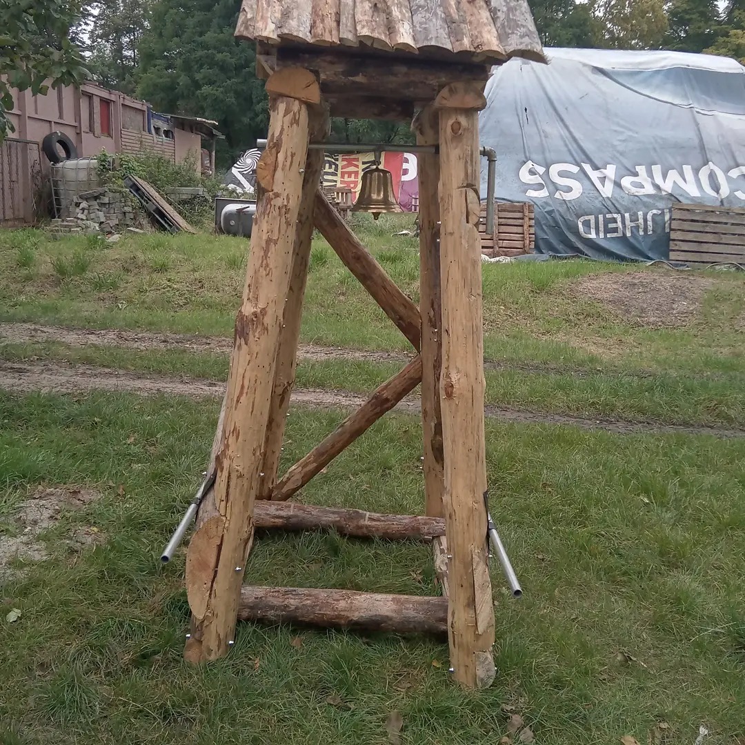 Drewniana konstrukcja z dzwonem, wykonana z surowych bali, zadaszona, na tle trawnika i zabudowań gospodarczych.