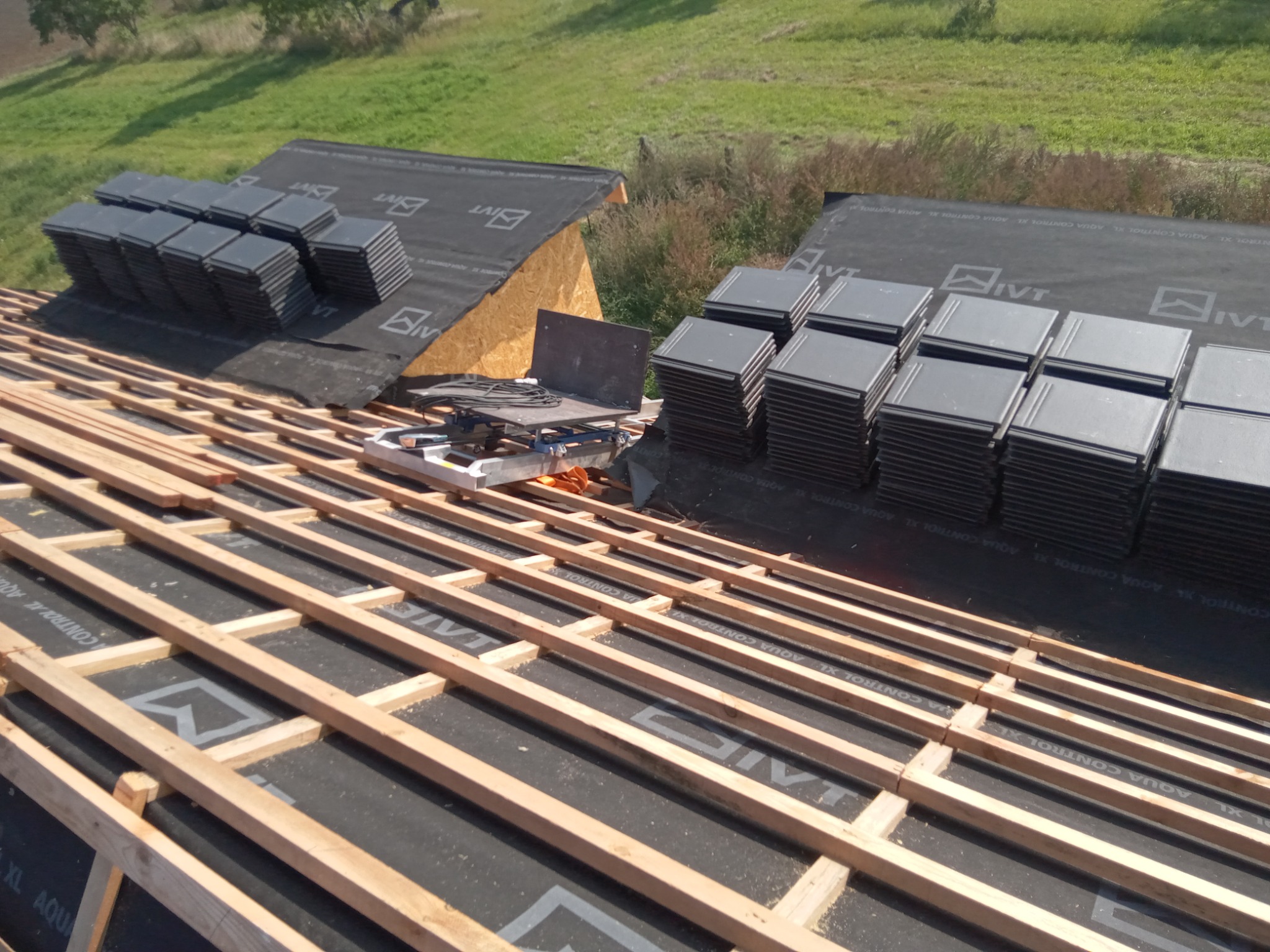 Konstrukcja dachu z łatami przygotowana do układania dachówki ceramicznej, widoczne stosy dachówek i membrana dachowa z nadrukowanym logo producenta.