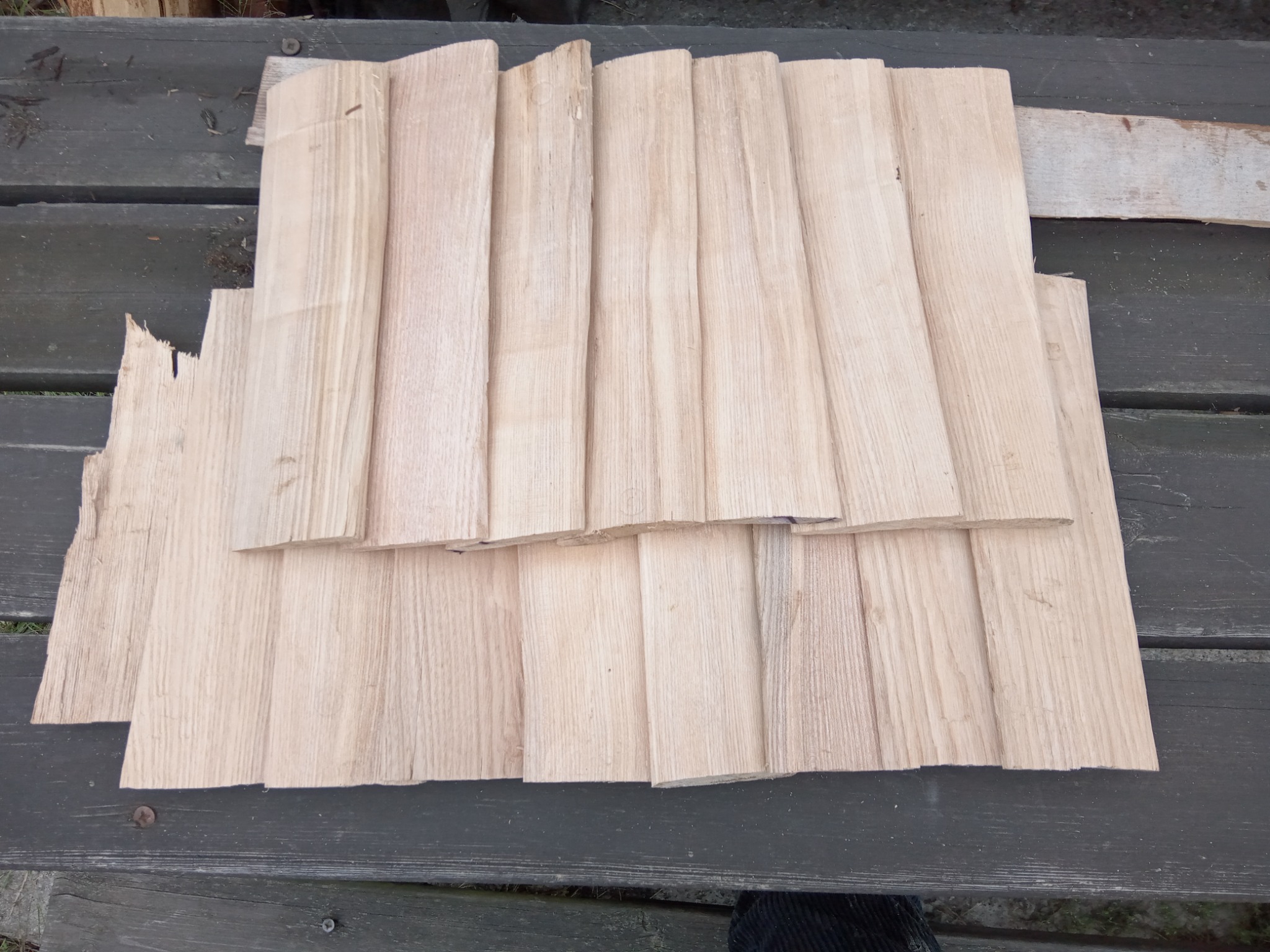 Kilkanaście drewnianych desek o jasnym odcieniu i widocznym usłojeniu, ułożonych na szarej, drewnianej powierzchni, niektóre z nich są przycięte nierówno.
