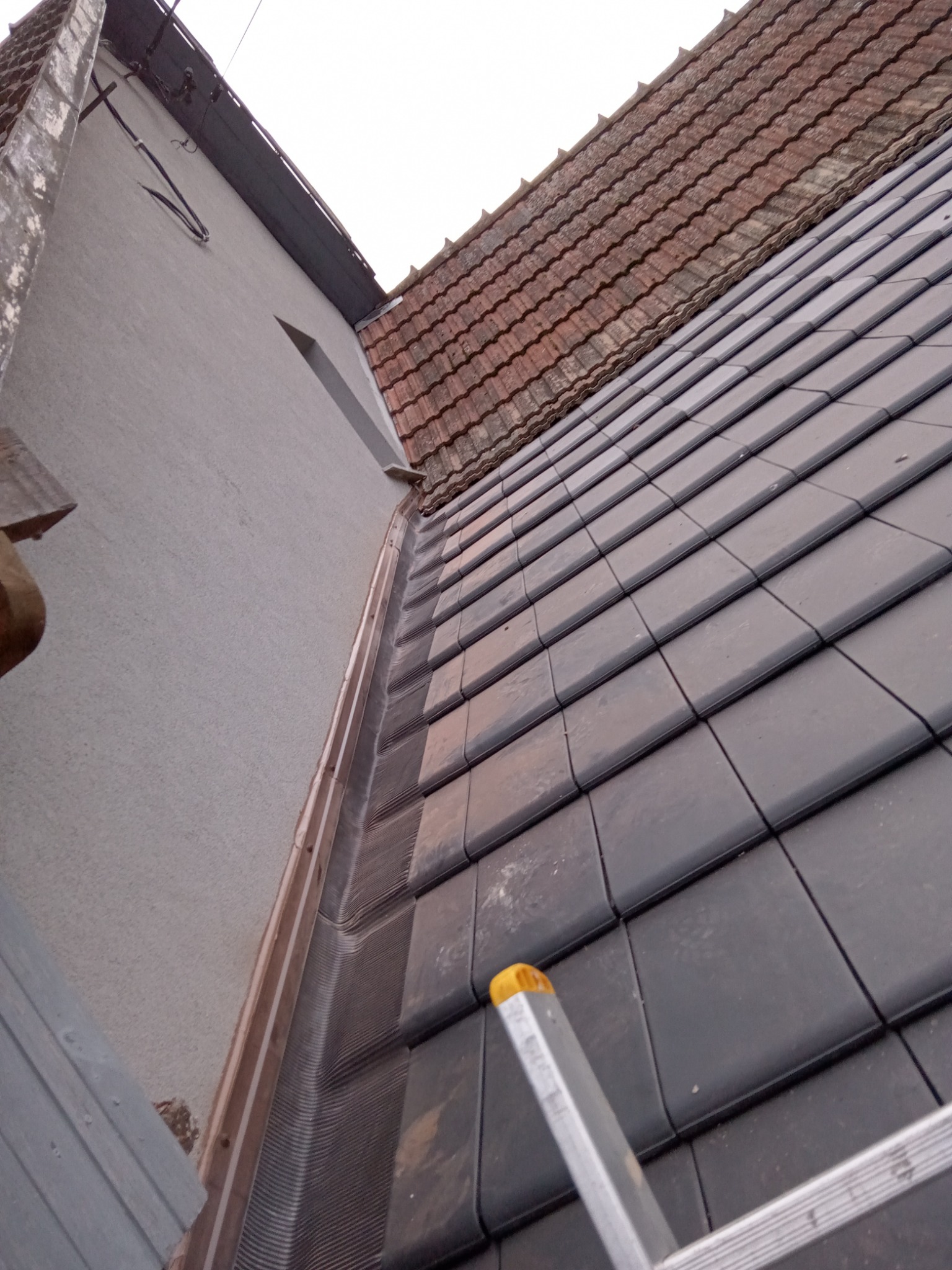 Fragment dachu z nową, grafitową dachówką ceramiczną i elementem drabiny w kadrze, widok z dołu na tle jasnej elewacji budynku.