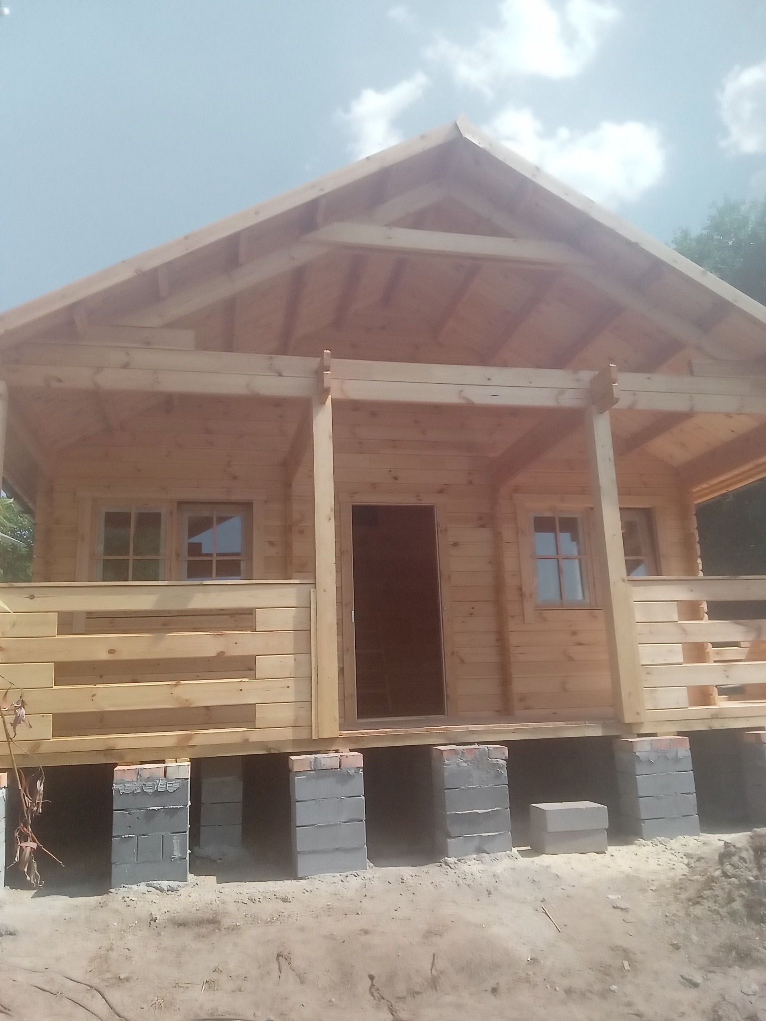 Surowy drewniany domek letniskowy w trakcie budowy, posadowiony na betonowych bloczkach, widoczne okna z szybami i zadaszony taras.
