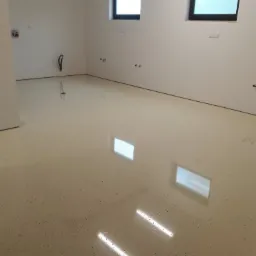 Jasna, błyszcząca posadzka żywiczna z delikatnym wzorem w przestronnym wnętrzu. Widoczne okna z czarnymi ramami i instalacje na ścianach.