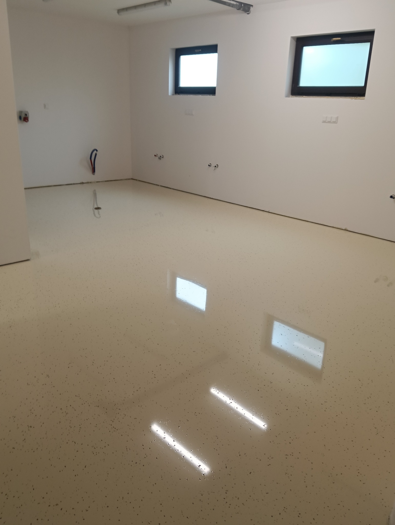 Jasna, błyszcząca posadzka żywiczna z delikatnym wzorem w przestronnym wnętrzu. Widoczne okna z czarnymi ramami i instalacje na ścianach.