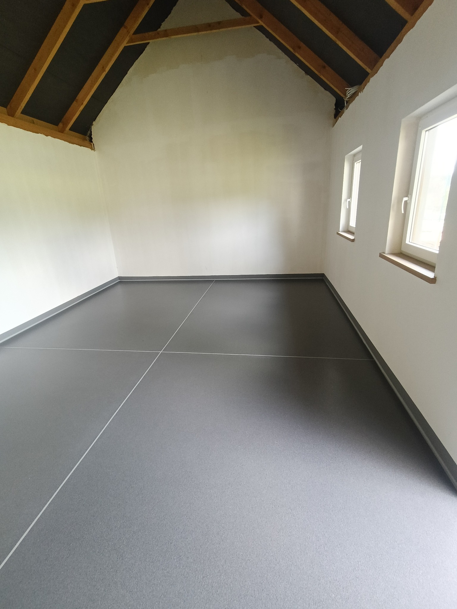 Szara, gładka podłoga z żywicy, przecięta białymi liniami, w jasnym pomieszczeniu z oknami i drewnianą konstrukcją dachu. Wykończone listwy przypodłogowe.