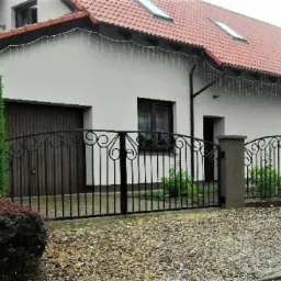 Czarna, metalowa brama wjazdowa z dekoracyjnymi elementami na tle domu z brązową bramą garażową i czerwonym dachem, oświetlona girlandą świetlną.