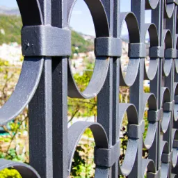 Żelazne ogrodzenie z dekoracyjnymi łukami na tle zieleni i zabudowań miejskich, widoczne detale metalowej konstrukcji i kamiennego słupka.