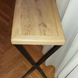 Blat dębowy stolika na metalowych nogach w kształcie litery X, widoczne słoje drewna i parkiet w jodełkę na podłodze.