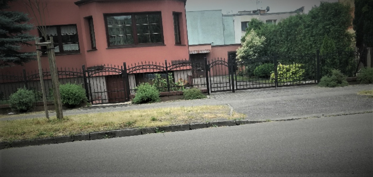 Kuta, czarna metalowa brama wjazdowa z ozdobnymi łukami, prowadząca do posesji z ceglanym budynkiem i zielenią, widok z ulicy.