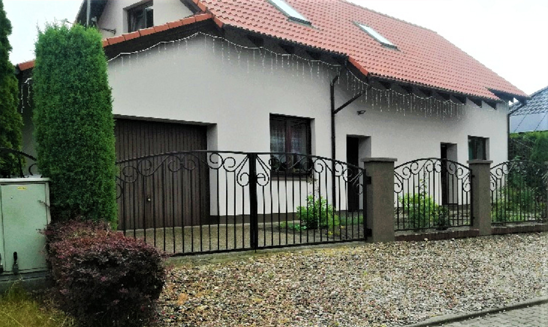 Czarna, metalowa brama wjazdowa z dekoracyjnymi elementami na tle domu z brązową bramą garażową i czerwonym dachem, oświetlona girlandą świetlną.