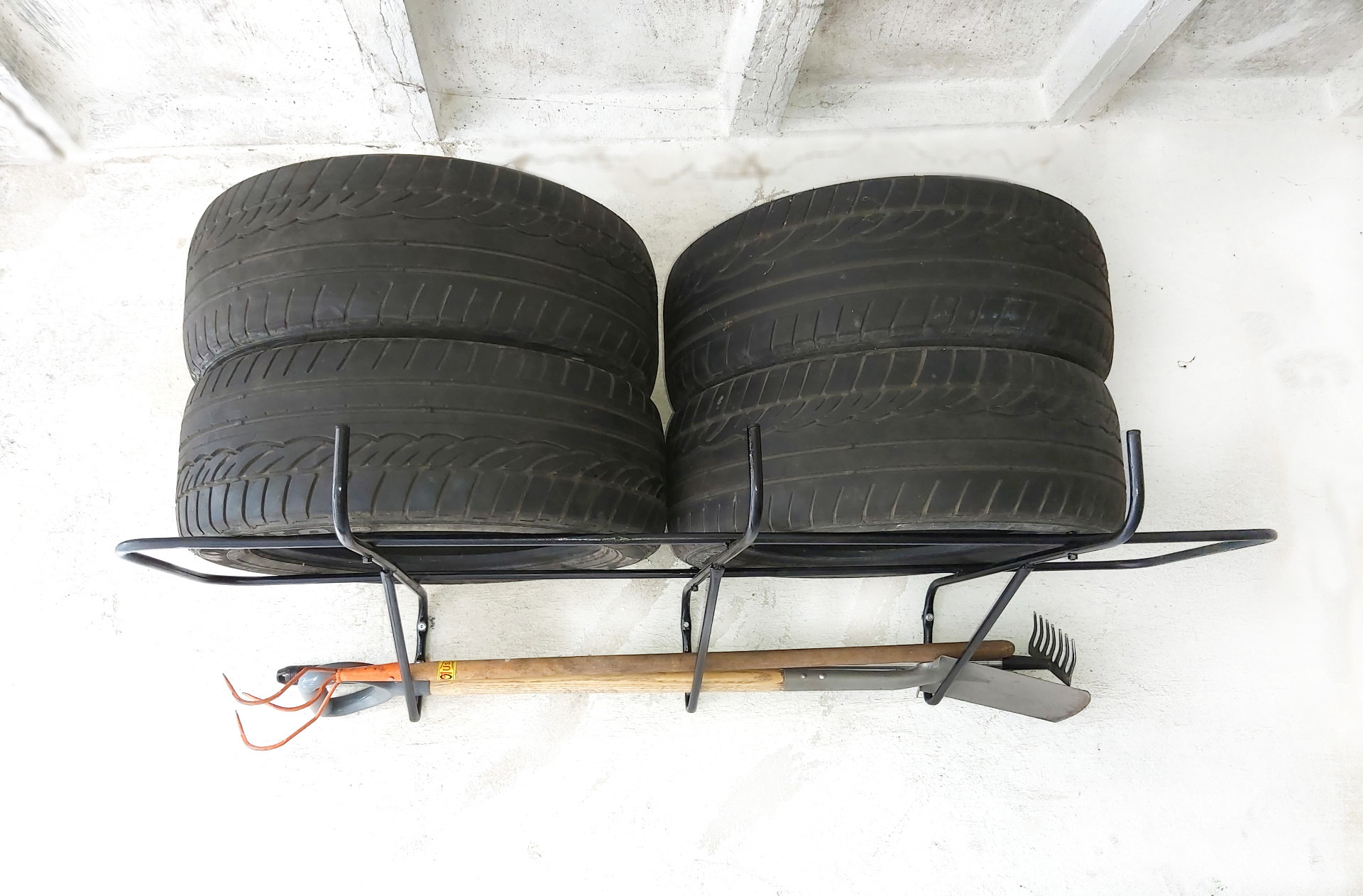 Dwie sterty opon samochodowych na metalowym regale przymocowanym do białej, lekko zabrudzonej ściany. Poniżej regału wiszą narzędzia ogrodnicze: grabie, szpadel i motyka.