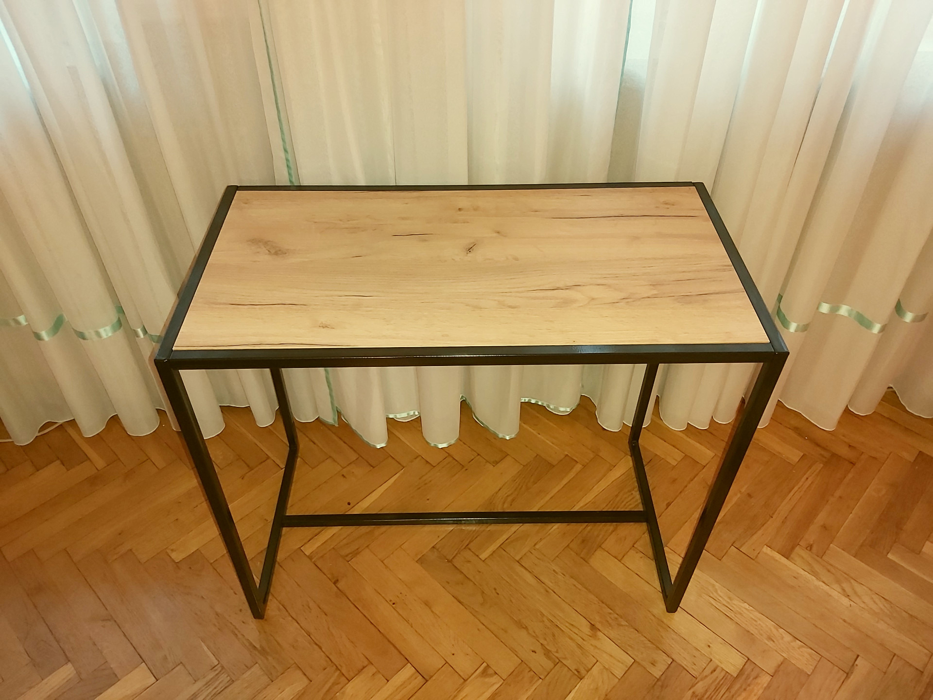 Stół z blatem imitującym jasne drewno i czarną, metalową ramą, na tle parkietu w jodełkę i białych zasłon z zielonym akcentem.
