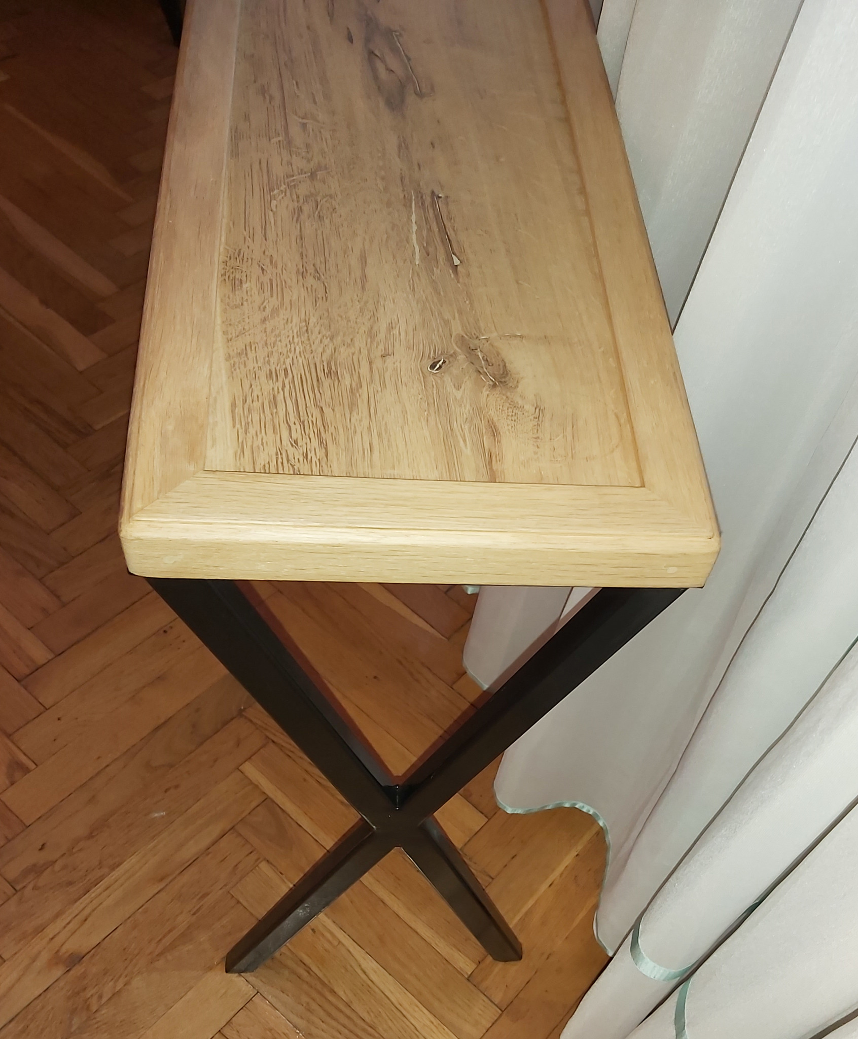 Blat dębowy stolika na metalowych nogach w kształcie litery X, widoczne słoje drewna i parkiet w jodełkę na podłodze.
