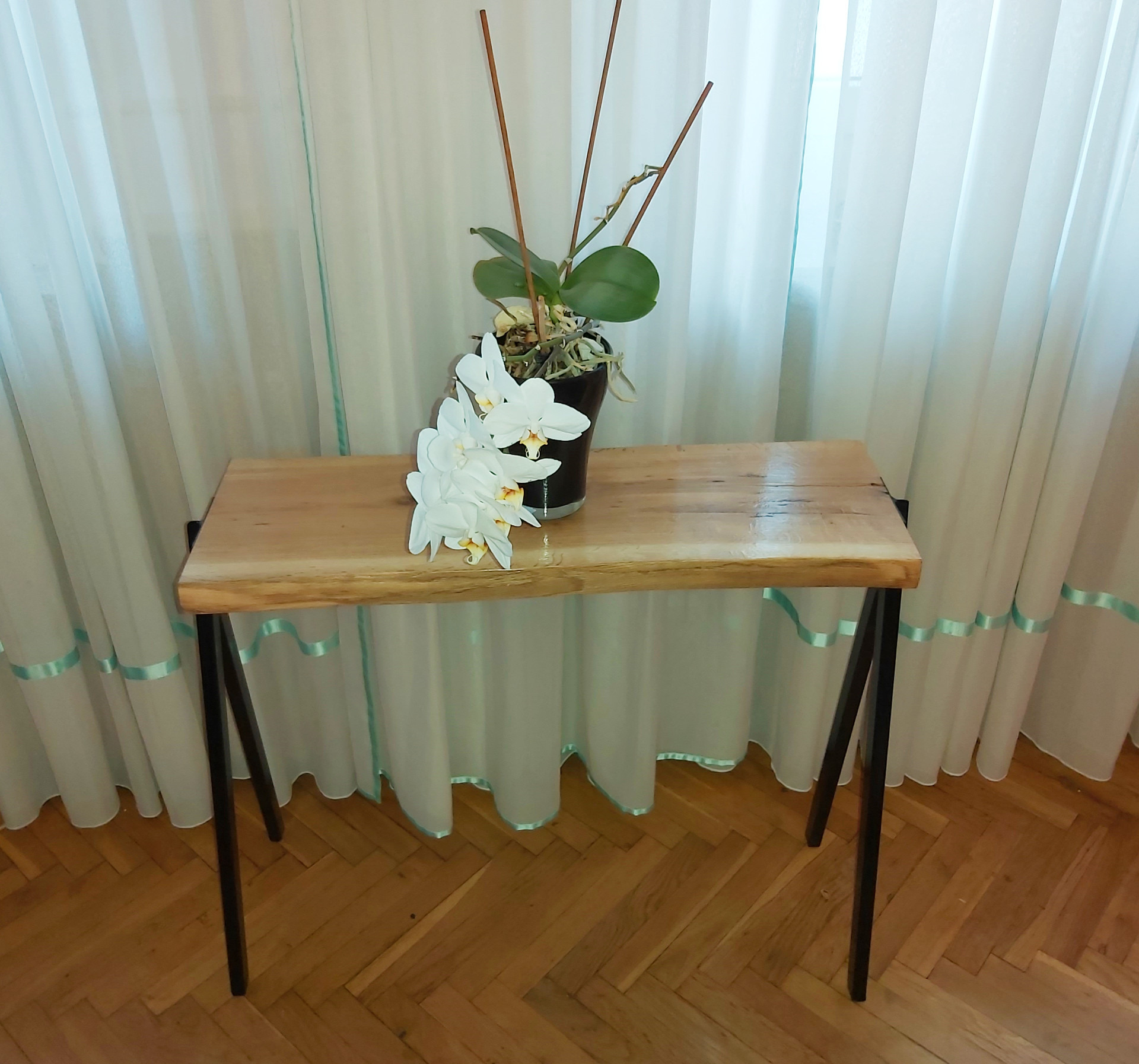Stolik kawowy z blatem z surowego drewna, czarne metalowe nogi, na blacie doniczka z białą orchideą, w tle białe zasłony z turkusową lamówką, parkiet w jodełkę.