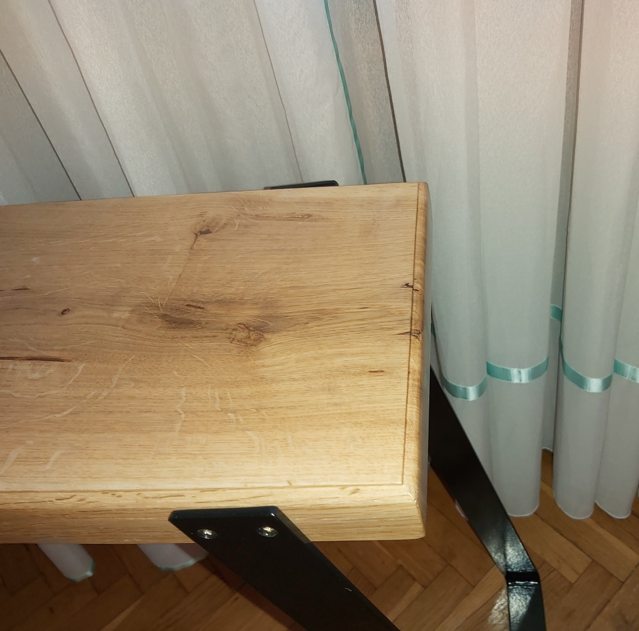 Blat dębowy na czarnych, metalowych nogach, widoczne słoje drewna i krawędź blatu, w tle jasne zasłony z turkusową wstążką.