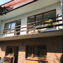 Dwupiętrowy dom z ceglaną elewacją na parterze i ocieplonym poddaszem, z drewnianą balustradą balkonu i widocznymi materiałami budowlanymi