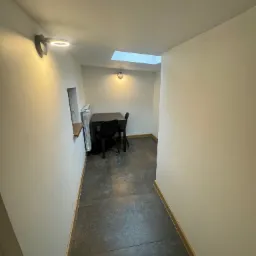 Wnętrze korytarza z szarymi płytkami podłogowymi, białymi ścianami oświetlonymi lampami punktowymi, z widokiem na mały pokój z oknem dachowym, czarnym stołem i krzesłami oraz grzejnikiem.