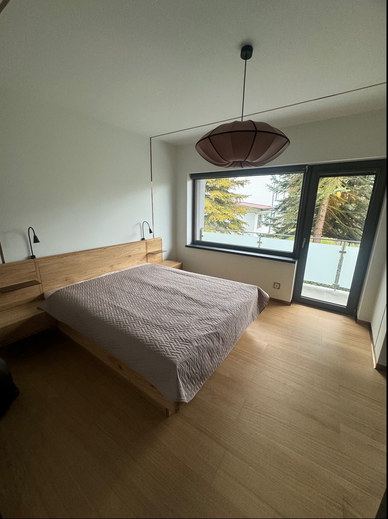 Jasne wnętrze sypialni z drewnianą podłogą i łóżkiem z wezgłowiem, lampkami nocnymi oraz widokiem na zieleń zza okna balkonowego.