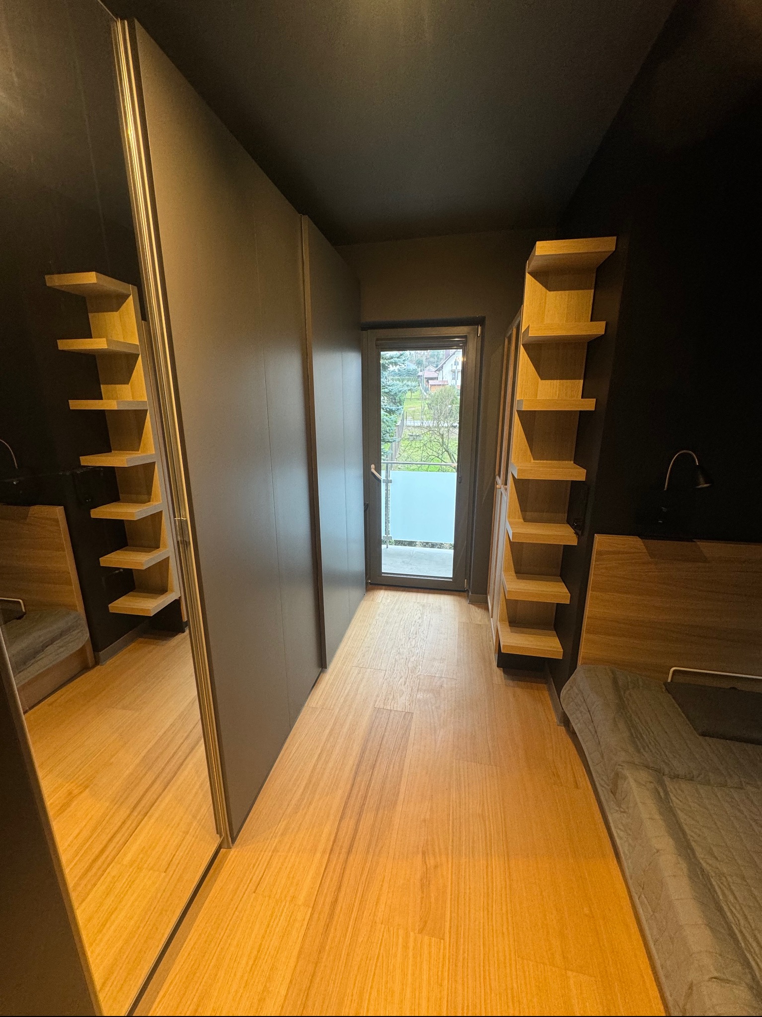 Nowoczesna sypialnia z ciemnymi ścianami, drewnianą podłogą i zabudową szafy z lustrem, widok na balkon przez przeszklone drzwi.