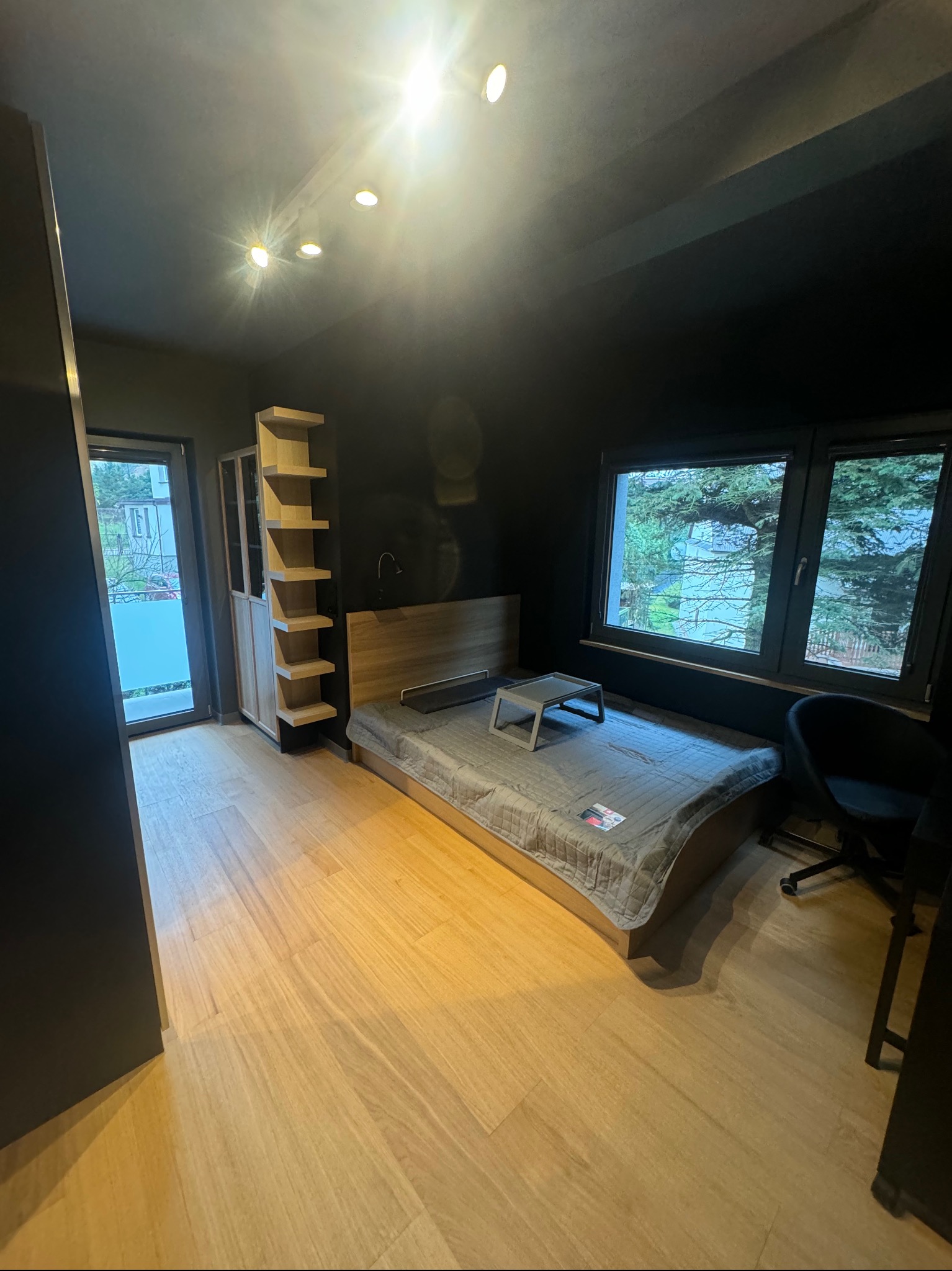 Pokój z ciemnymi ścianami, jasną drewnianą podłogą i łóżkiem z szarą narzutą, widoczne okno z widokiem na zieleń i regał z jasnego drewna