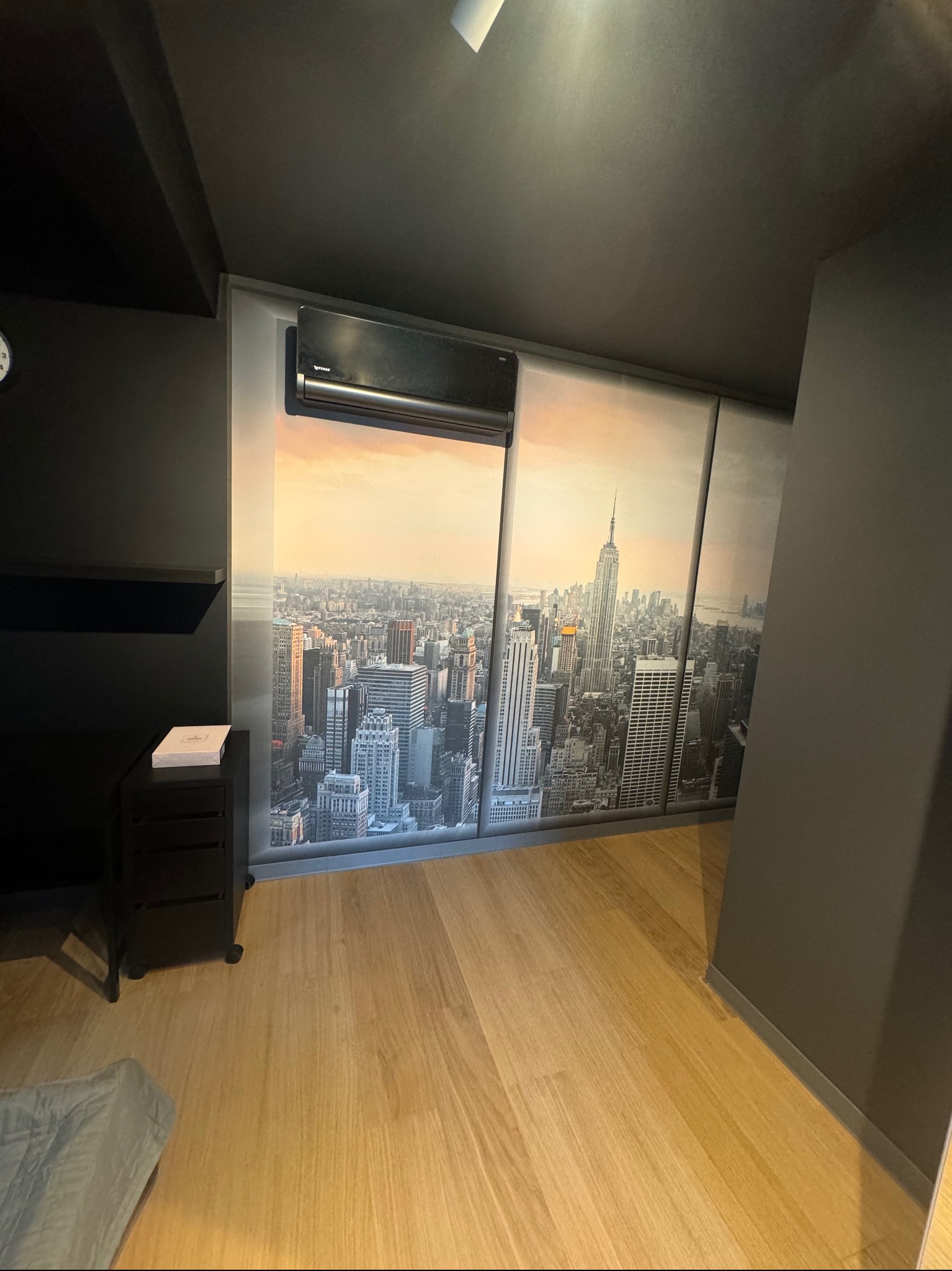 Wnętrze pokoju z panelami podłogowymi i fototapetą przedstawiającą panoramę Nowego Jorku z Empire State Building, klimatyzacja na ścianie, czarne meble.