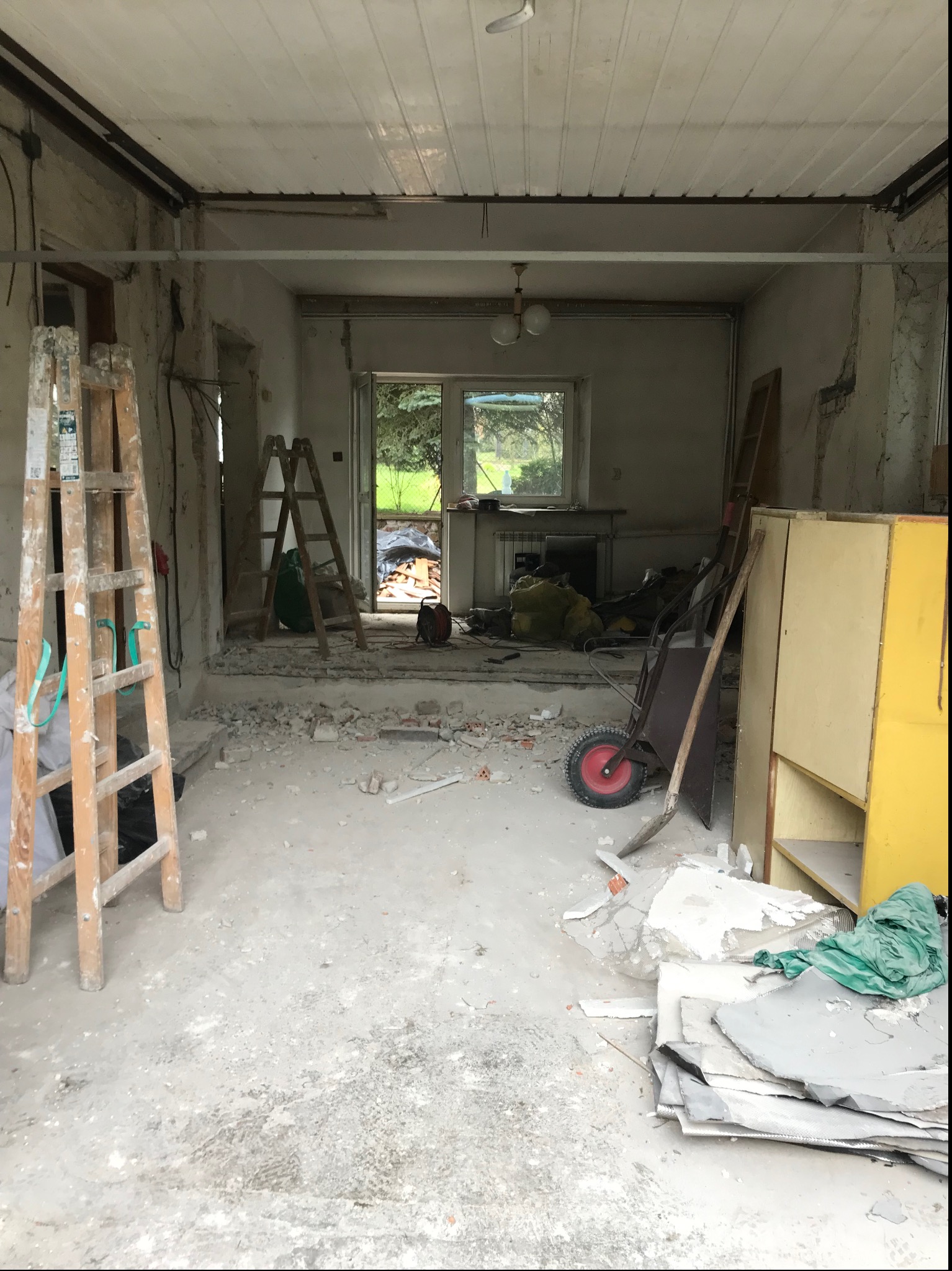Wnętrze w trakcie gruntownego remontu: widoczne dwie drabiny, gruz na podłodze, odsłonięte ściany, taczka i szafka w tle, okno z widokiem na zieleń.