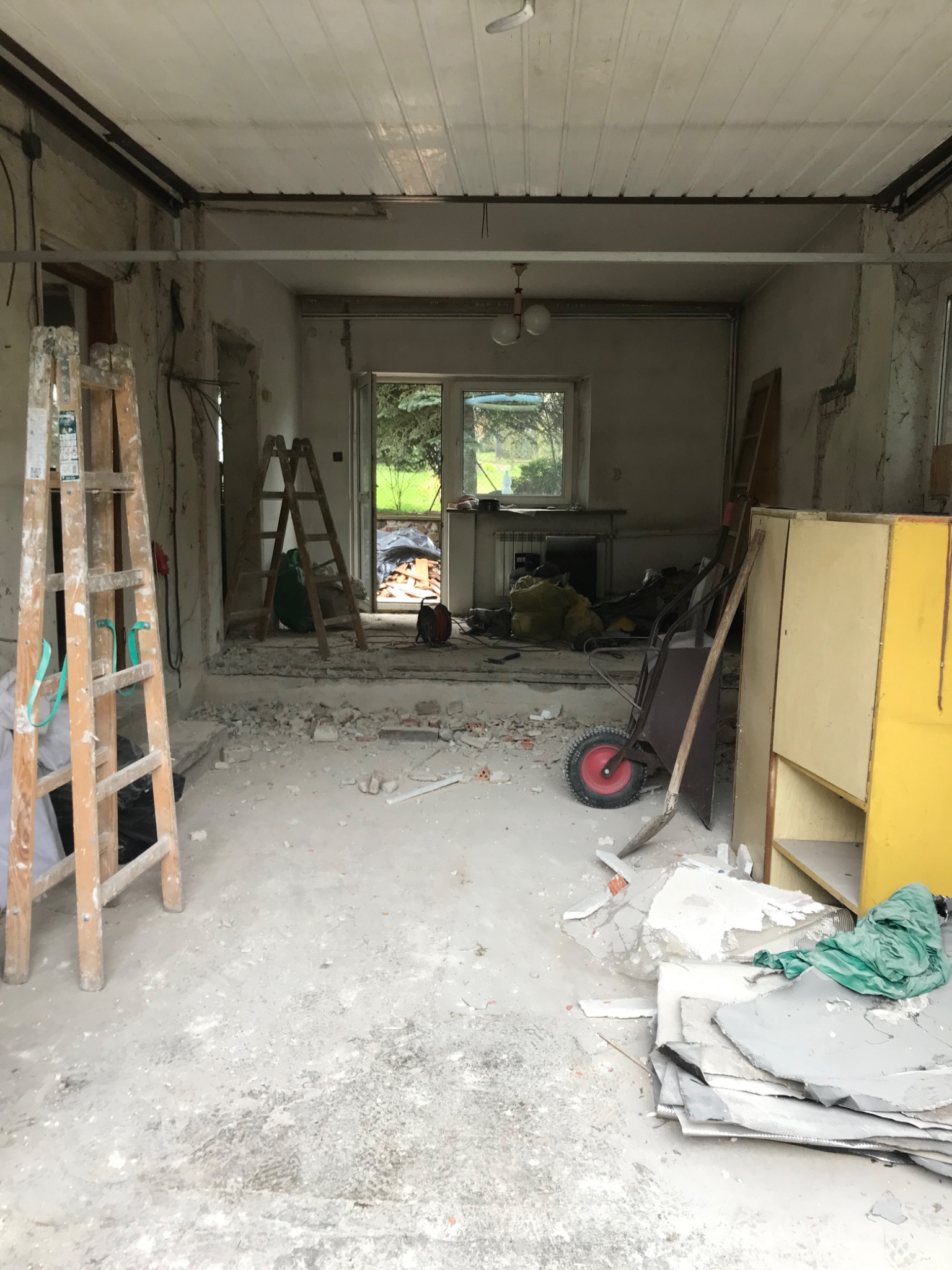 Wnętrze w trakcie demontażu: widoczne dwie drabiny drewniane, gruz na podłodze, taczka, fragmenty płyt i odsłonięta instalacja elektryczna pod sufitem.