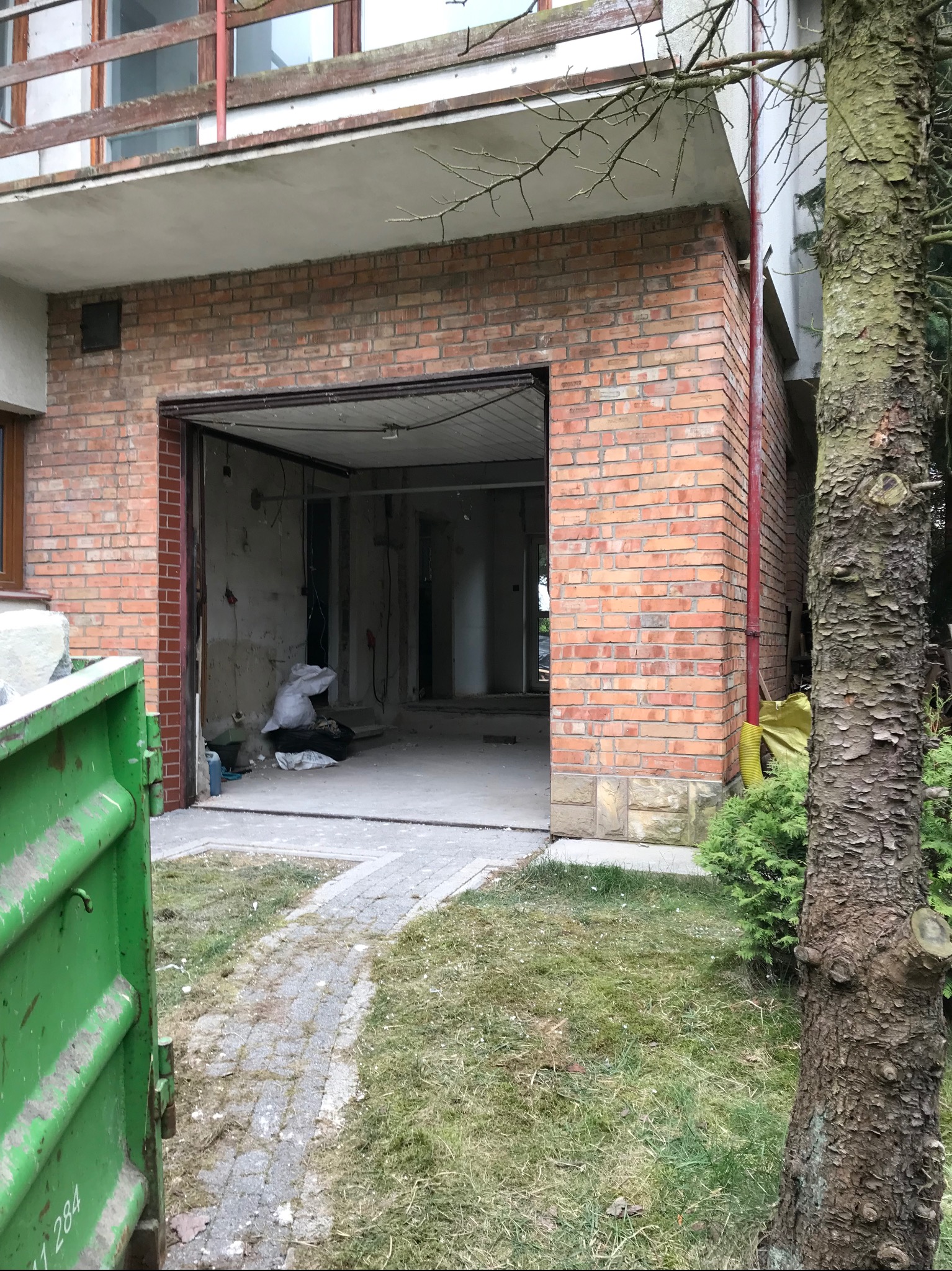 Ceglana dobudówka garażu w trakcie remontu, widoczny otwarty wjazd, worki z gruzem w środku i zielony kontener na odpady na zewnątrz.